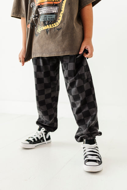 A person wearing loose black and gray checkered pants, a brown graphic t-shirt, and black-and-white sneakers, standing against a clean white studio background.