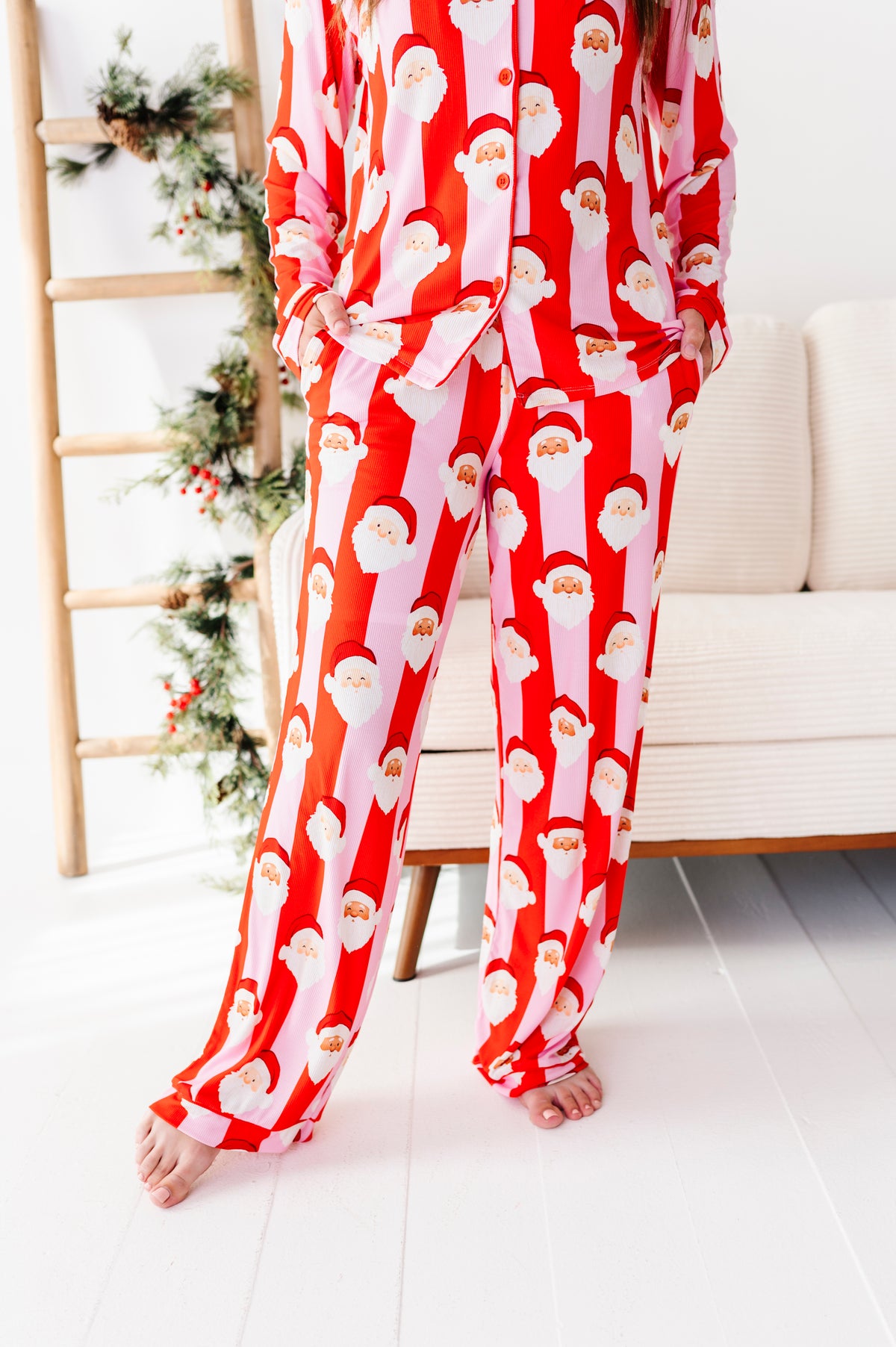 A person wearing a red and white Santa Claus patterned bamboo pajama set, standing barefoot in a bright room with a sofa and holiday ladder visible in the background.
