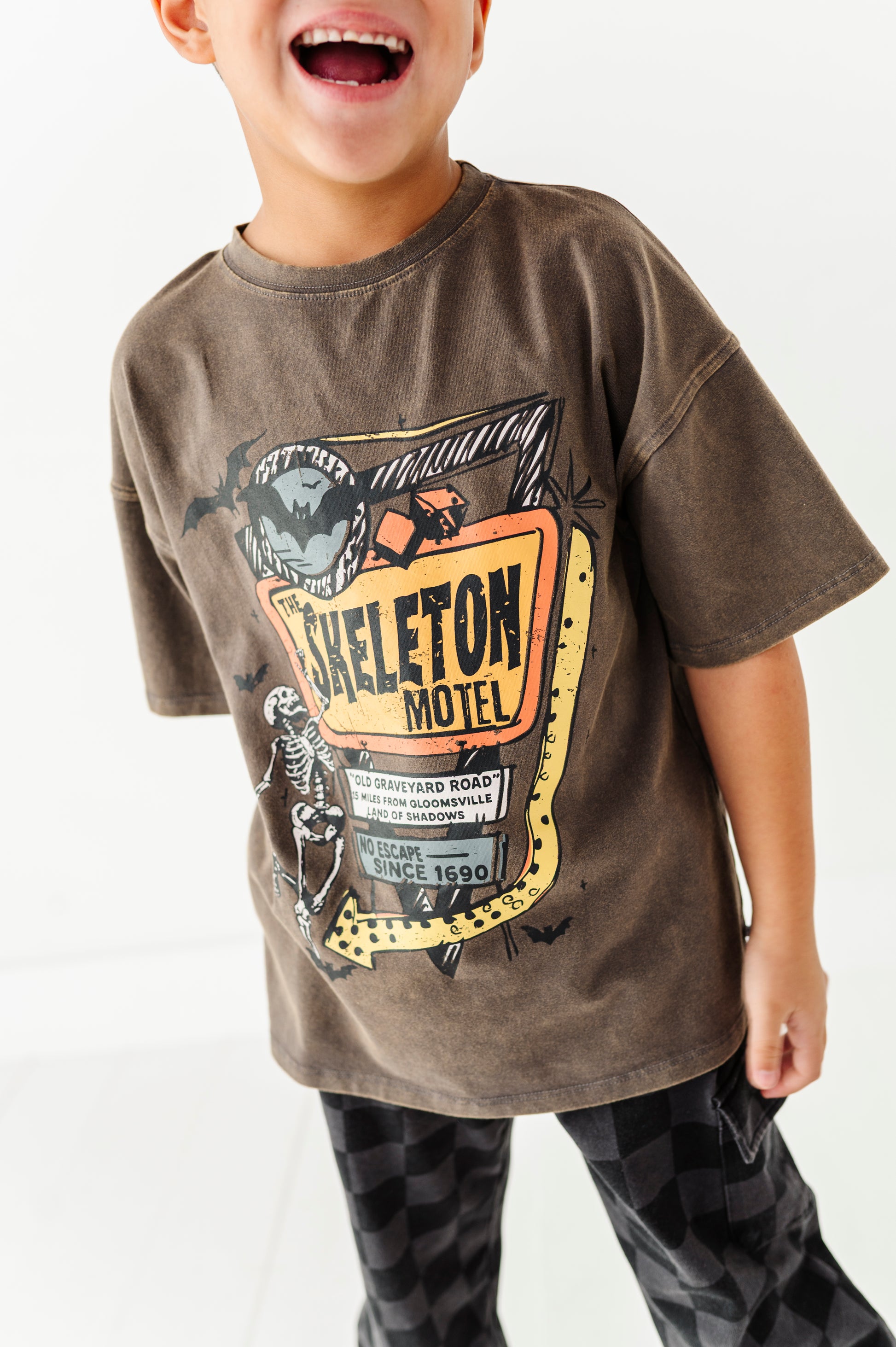 A child wearing a brown graphic t-shirt with a vintage 'Skeleton Motel' print, paired with black and white checkered pants, against a plain white background.