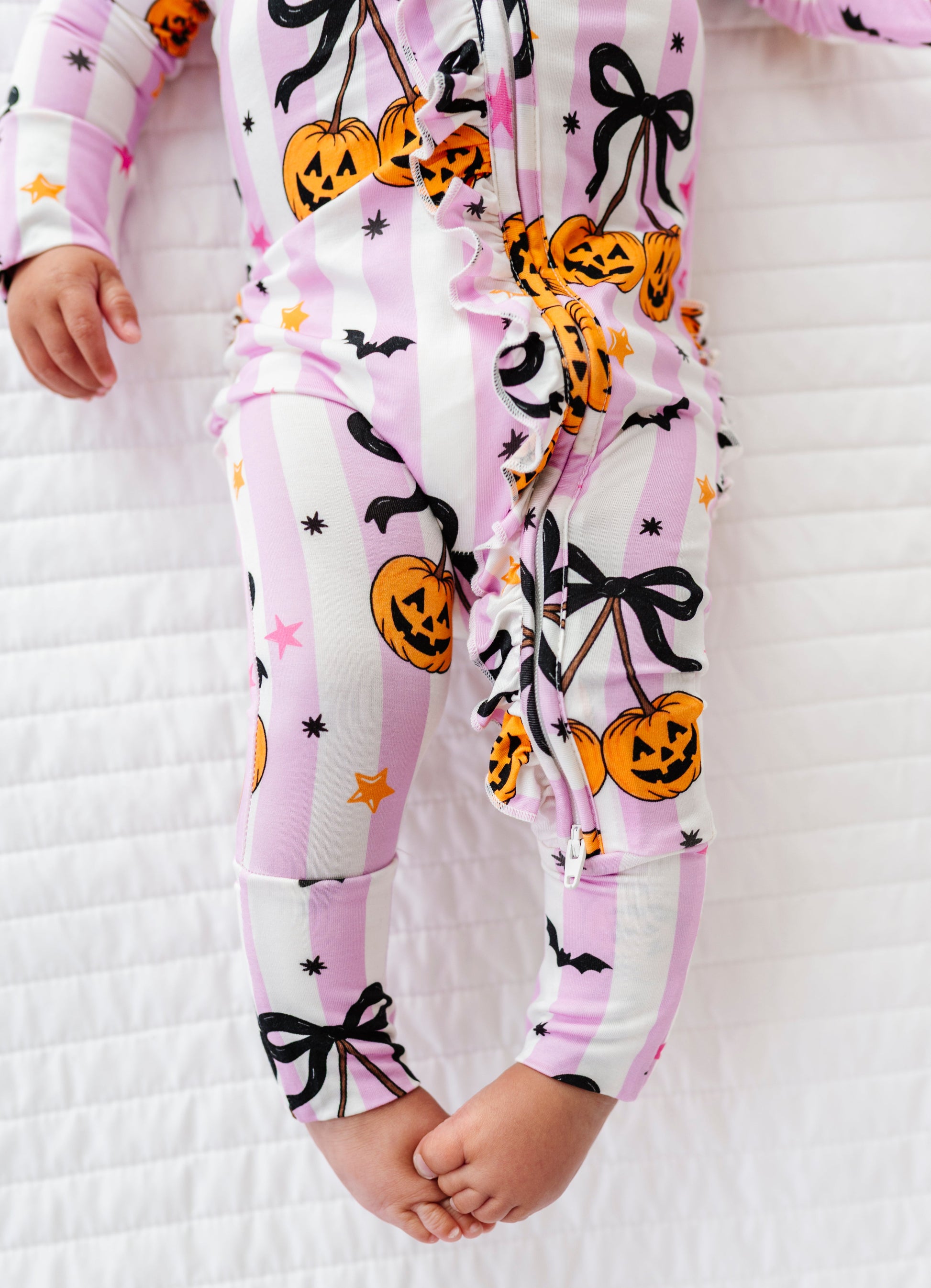 A baby wearing a pink and white striped Halloween-themed footless one-piece jumpsuit with jack-o'-lanterns, black bows, and stars, lying on a white quilted blanket.