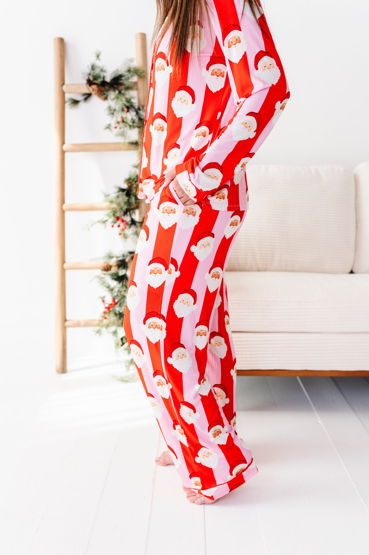 A person wearing a red and white Santa Claus patterned bamboo pajama set, standing in a bright room with a white sofa and a wooden ladder decorated with greenery.