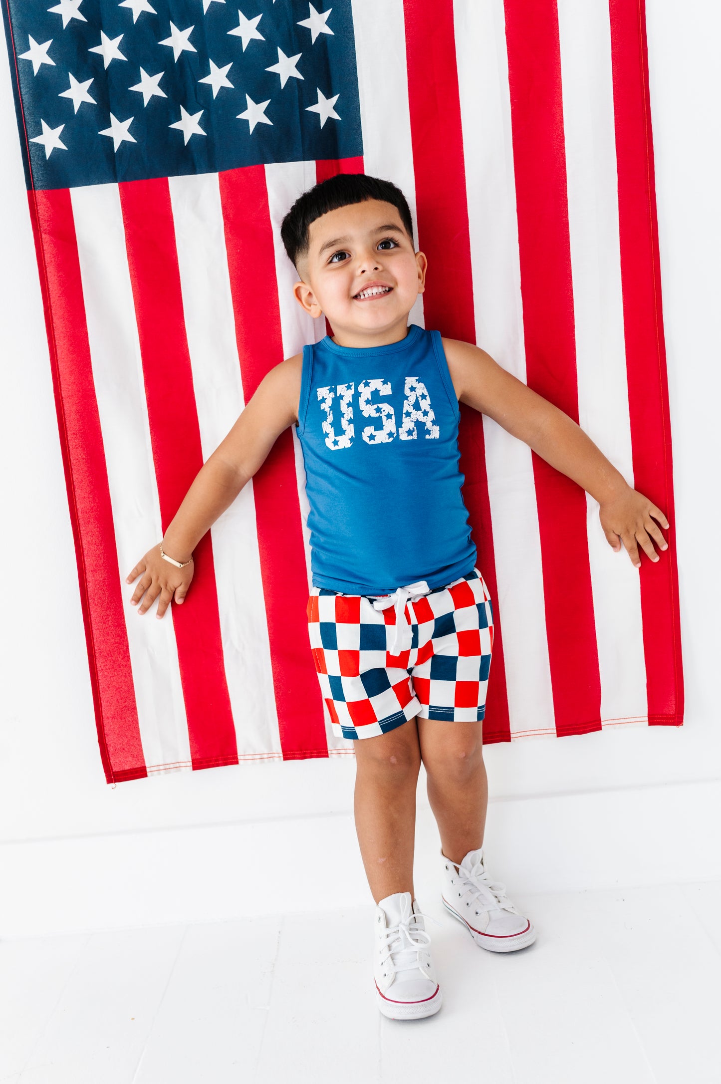 Child wearing a blue 'USA' tank top and checkered shorts standing in front of an American flag.
