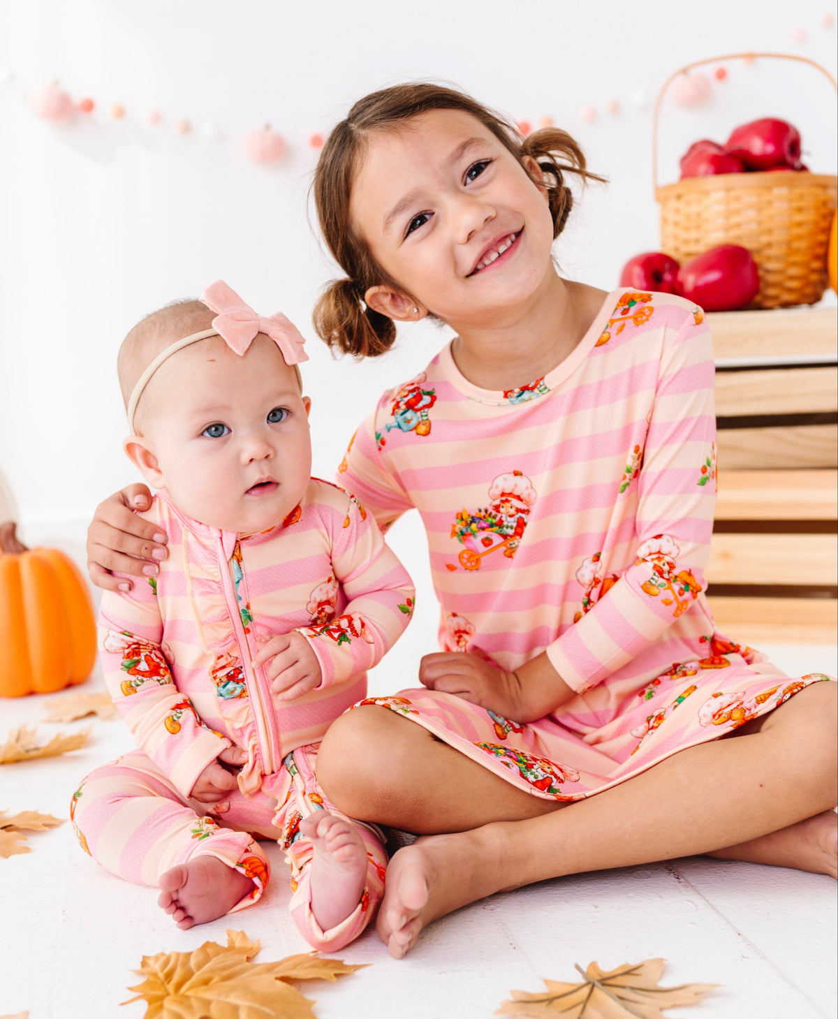 Two young children sitting on a white floor wearing matching pink striped pajamas with a colorful autumn leaf print, surrounded by fall-themed props including pumpkins, leaves, and baskets of red apples.