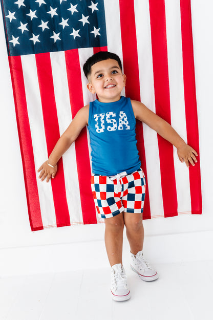 Child wearing a blue 'USA' tank top and checkered bamboo shorts standing in front of an American flag.