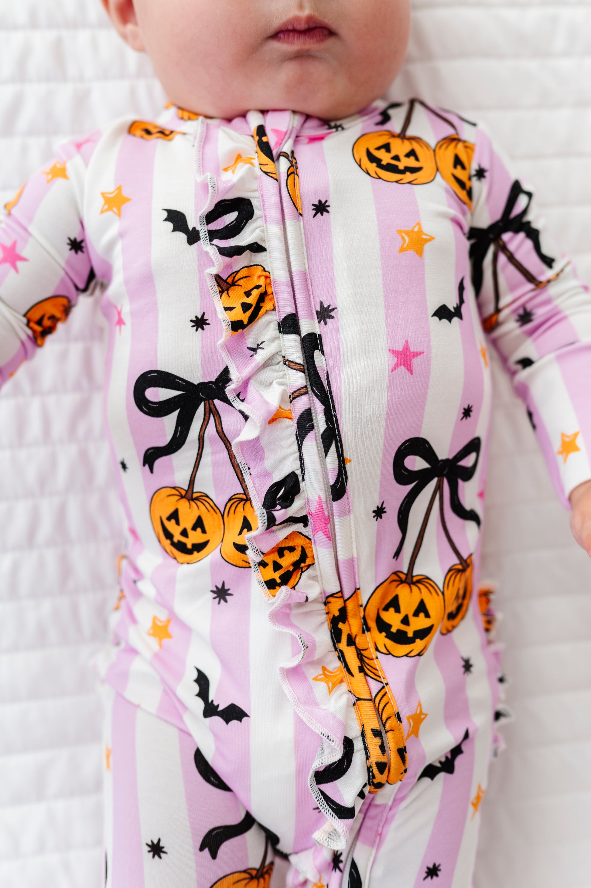 A close-up of a baby wearing a pink and white striped Halloween onesie with orange jack-o'-lanterns, black bows, bats, and stars, lying on a white textured blanket.