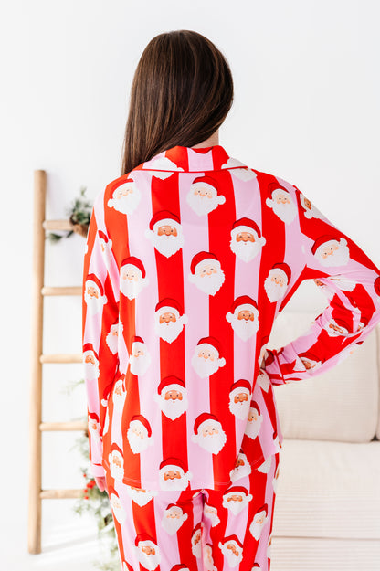 A woman viewed from behind wearing a red and pink bamboo pajama set with an all-over Santa Claus face print, standing in a bright indoor room.