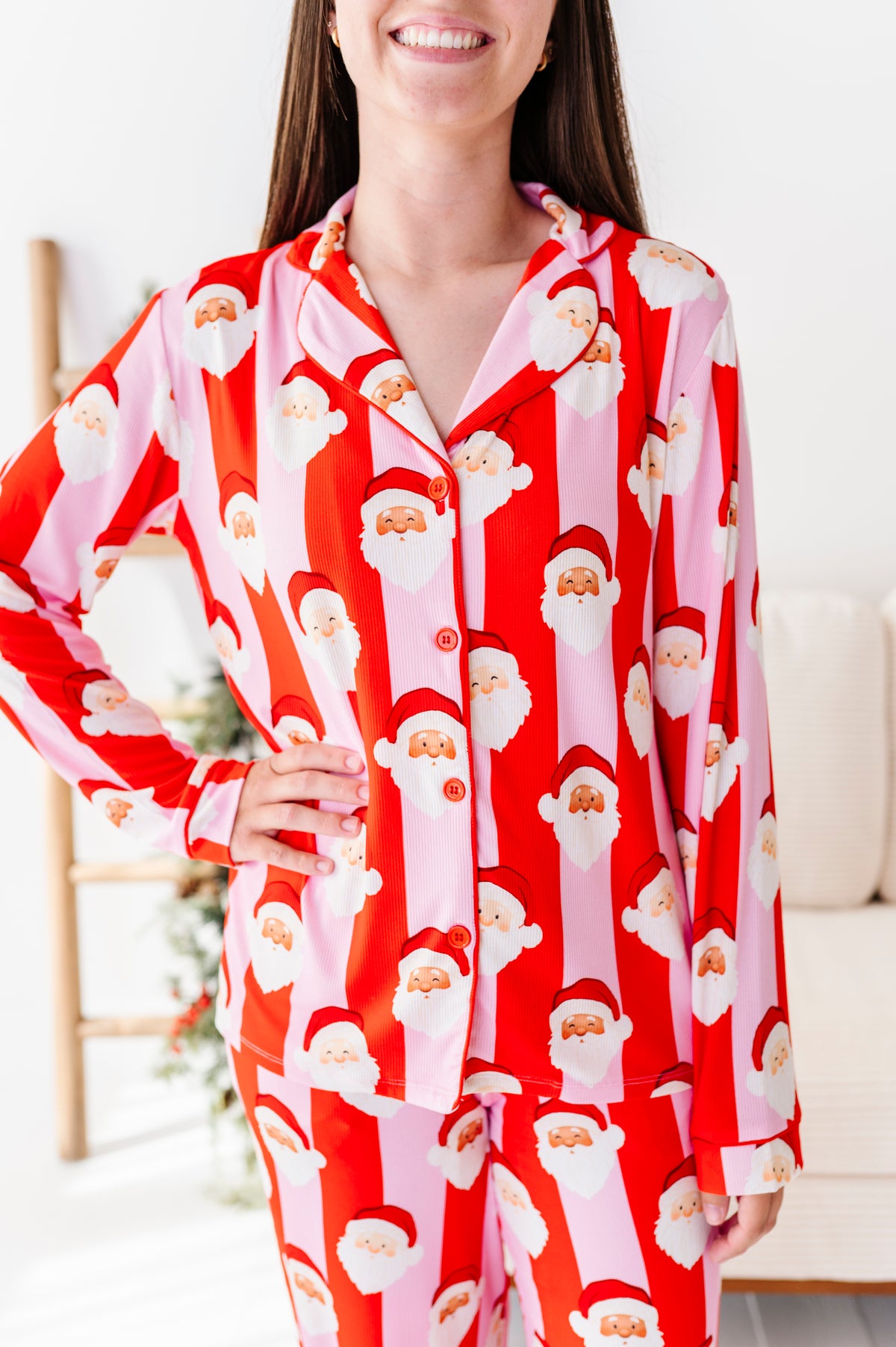 A woman wearing a red and pink bamboo pajama set with a repeating Santa Claus face pattern, standing indoors with a smiling expression.