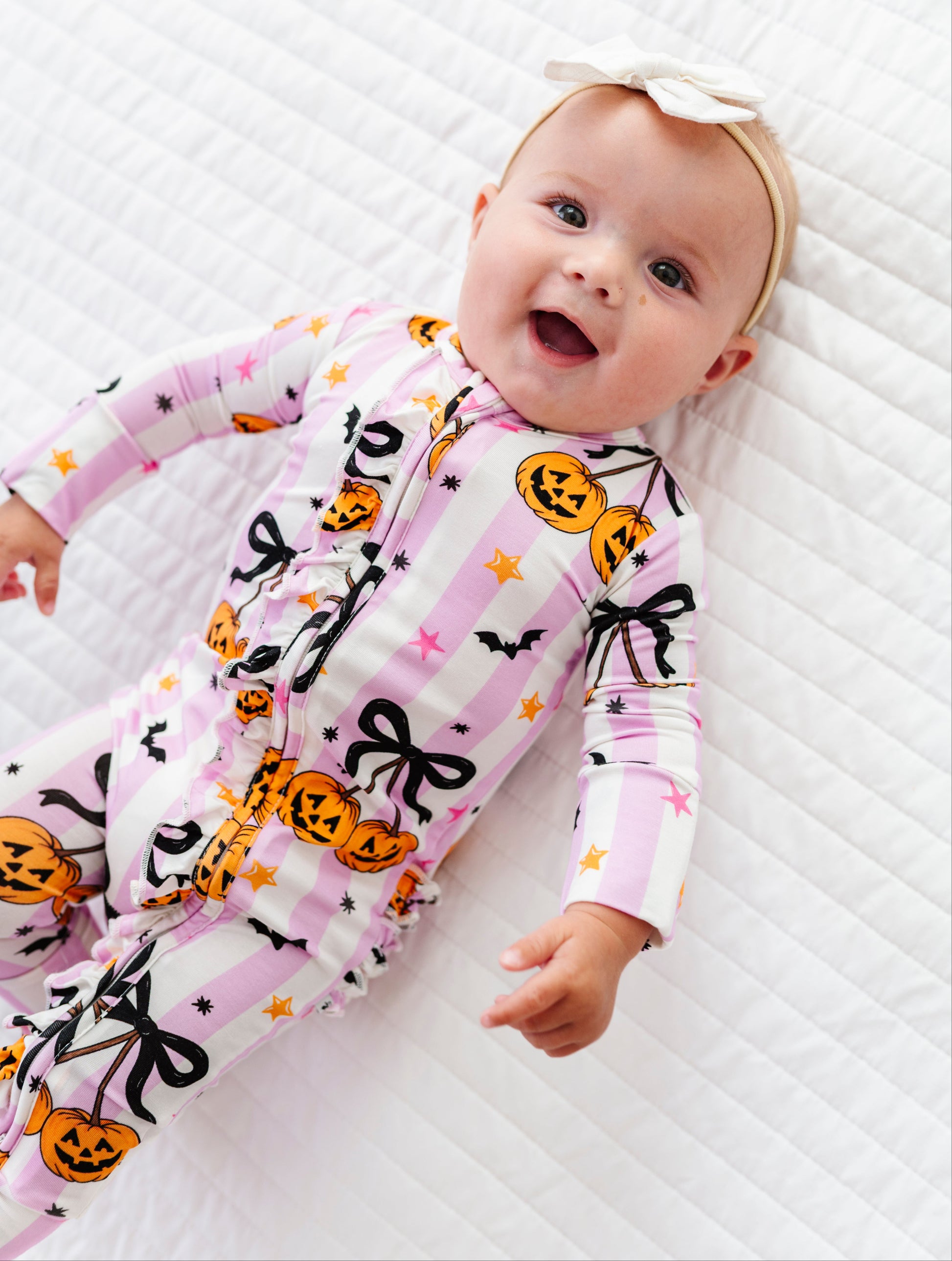A baby lying on a white quilted mattress wearing a pink and white striped Halloween-themed footless one-piece pajama with orange jack-o'-lanterns, black bows, and bats, along with a white bow headband.