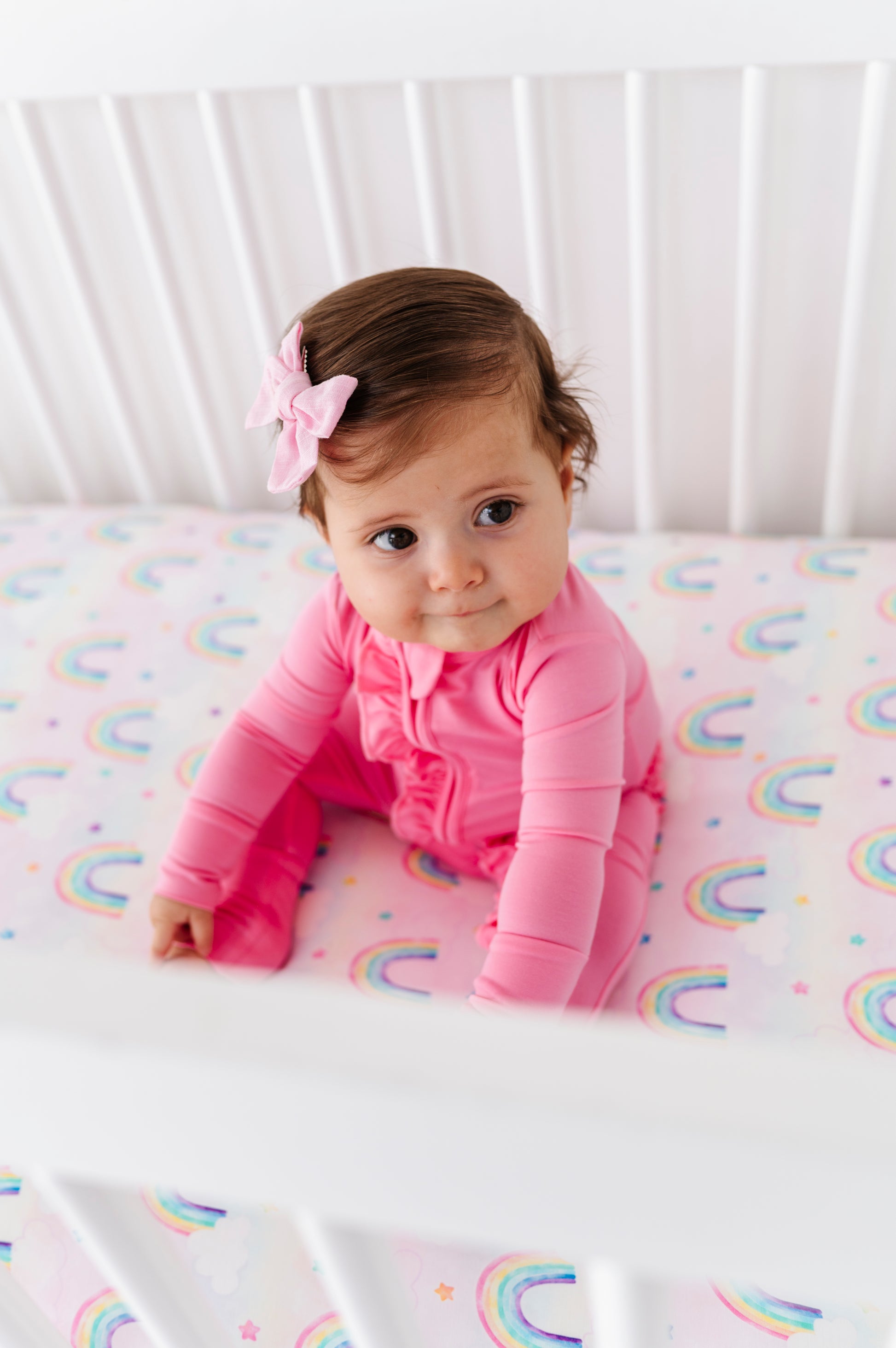 Baby in a pink bamboo zip romper sitting on a crib with rainbow patterned bamboo crib sheet.