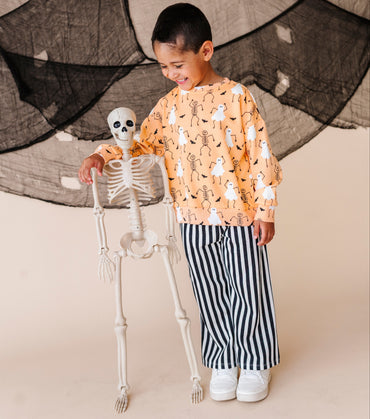 Child in Halloween-themed outfit with skeleton and ghost pattern, standing next to a skeleton decoration.