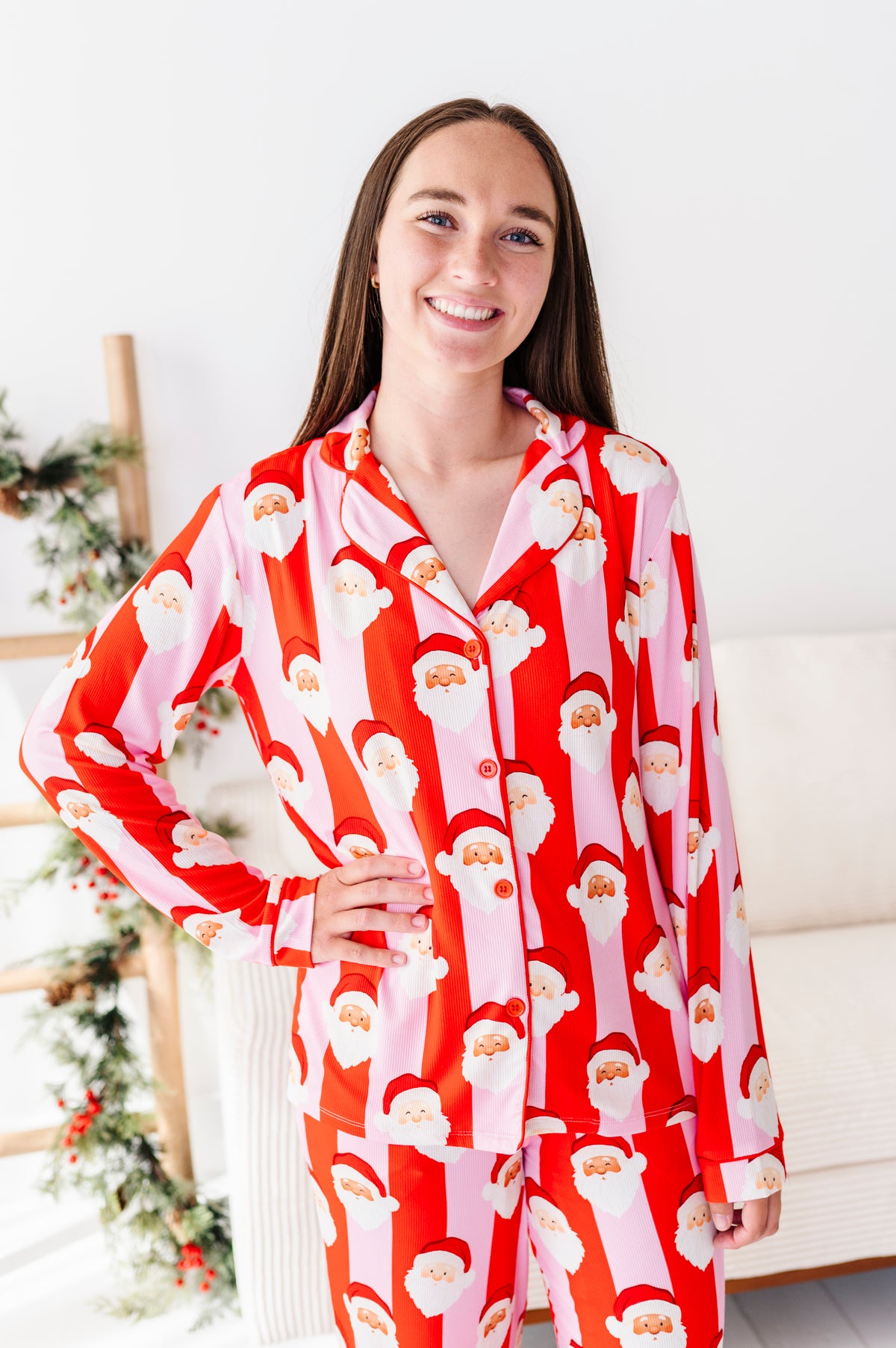 A woman wearing a red and pink bamboo pajama set with a repeating Santa Claus print, standing in a bright indoor room with a white couch and holiday decor in the background.