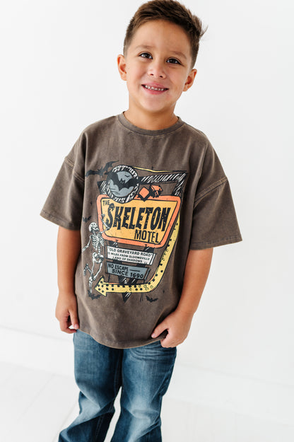 A young boy smiling and wearing a brown graphic t-shirt with a vintage 'Skeleton Motel' print, paired with blue jeans, against a plain white background.