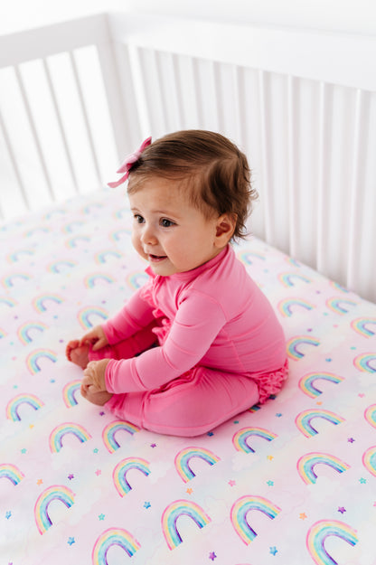 Baby wearing a bamboo convertible romper and sitting on a crib with a rainbow-patterned sheet