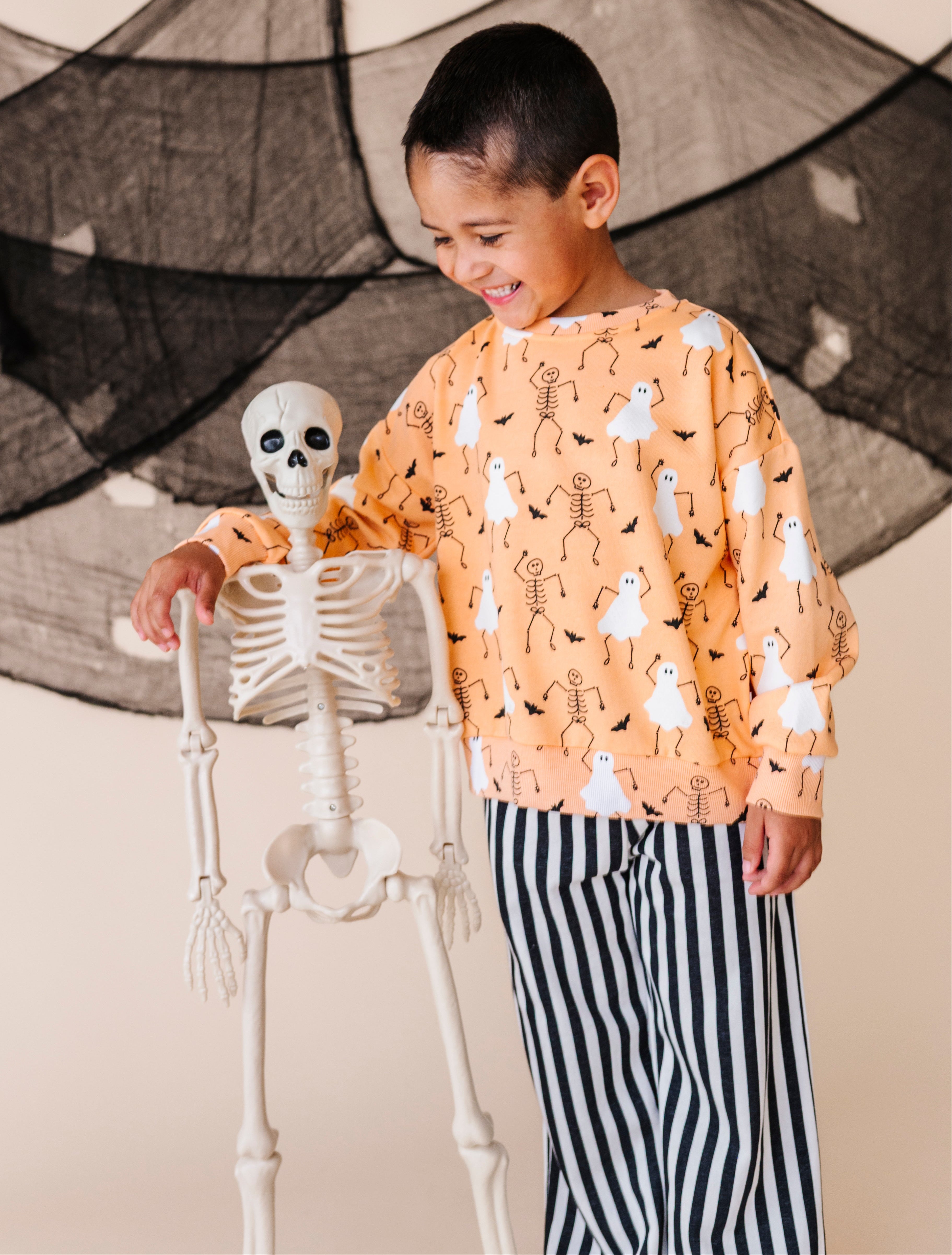 Child wearing a Halloween-themed sweatshirt with a skeleton and ghost design, standing next to a small skeleton figure.