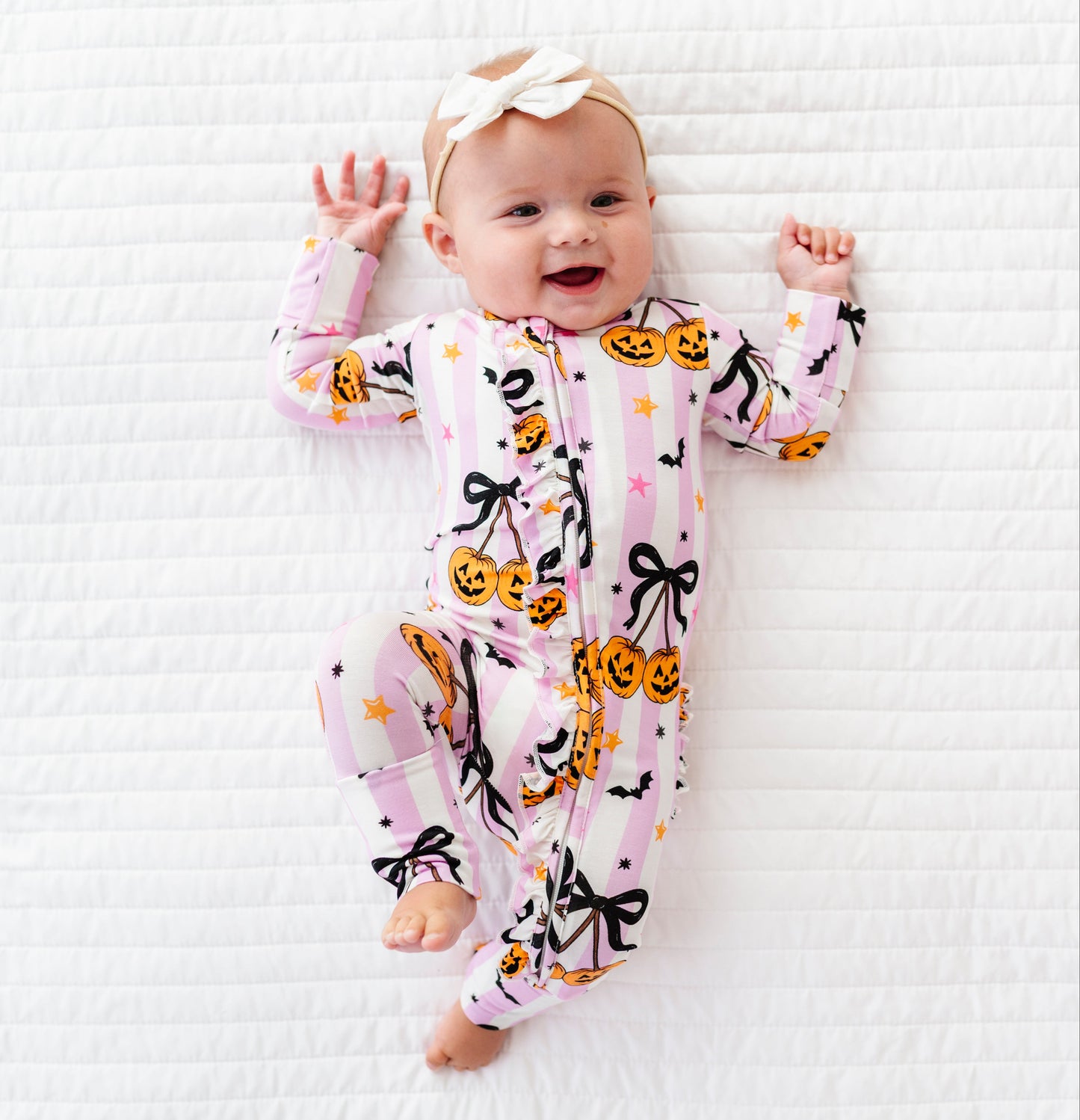 A baby lying on a white textured blanket, wearing a pink one-piece pajama with a Halloween print of pumpkins, black bows, and bats, along with a white bow headband.
