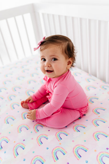 Baby wearing a bamboo zip romper and sitting on bamboo crib sheets featuring watercolor pastel rainbows 
