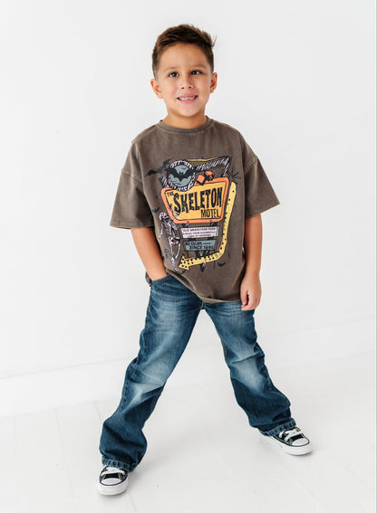 A young boy modeling a brown graphic t-shirt with a 'SKELETON ARMY' design, paired with blue jeans and black-and-white sneakers, standing against a white studio background.