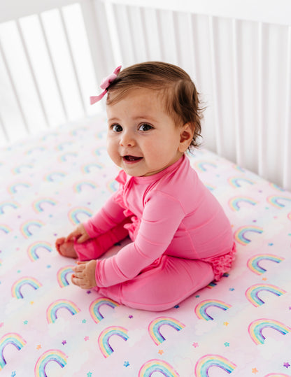 Baby sitting on a crib with a pink bamboo convertible romper and rainbow-patterned sheet.