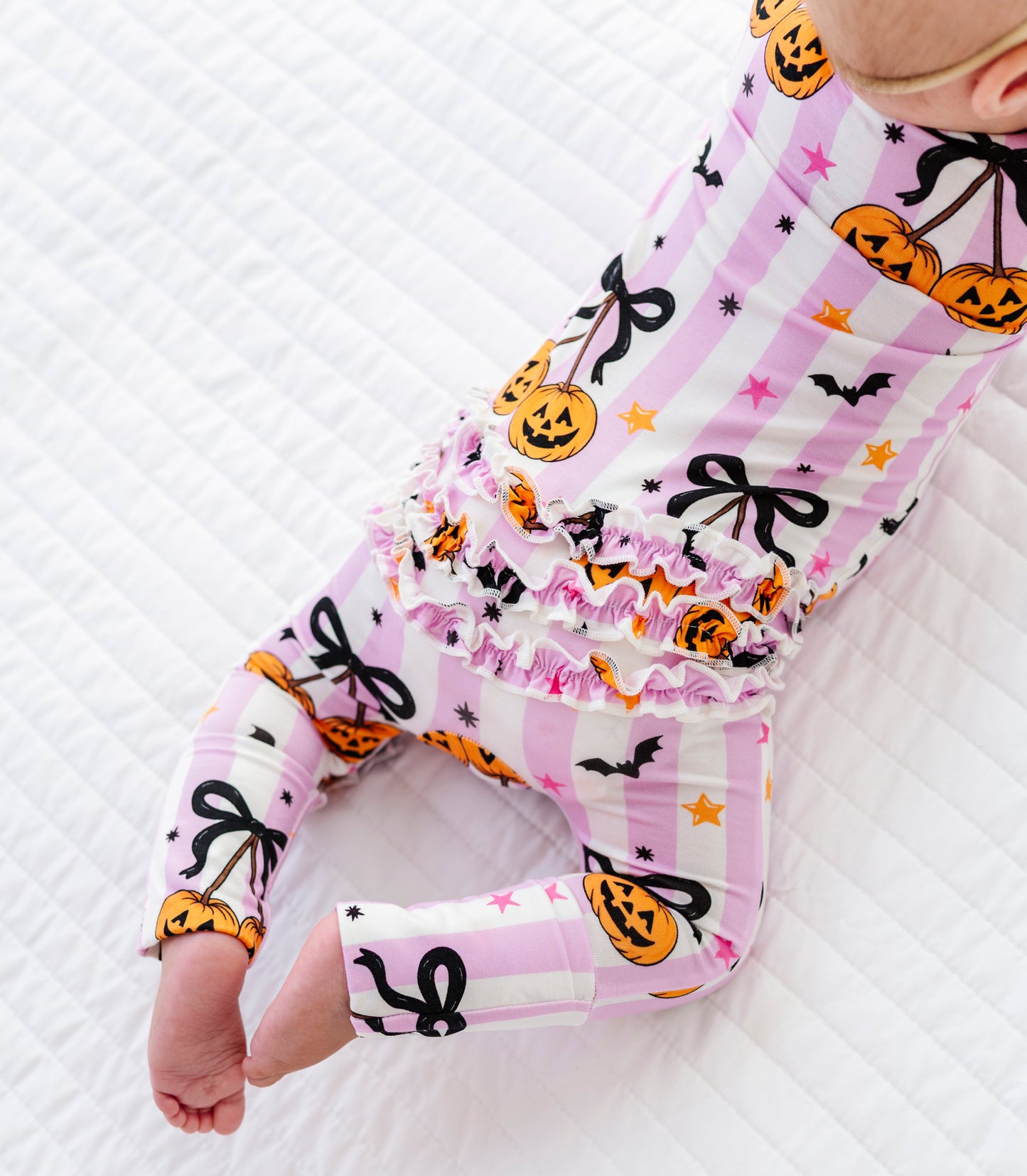 A baby wearing a pink and white striped long-sleeve romper with a Halloween print of pumpkins, black bows, and bats, lying on a white quilted blanket.