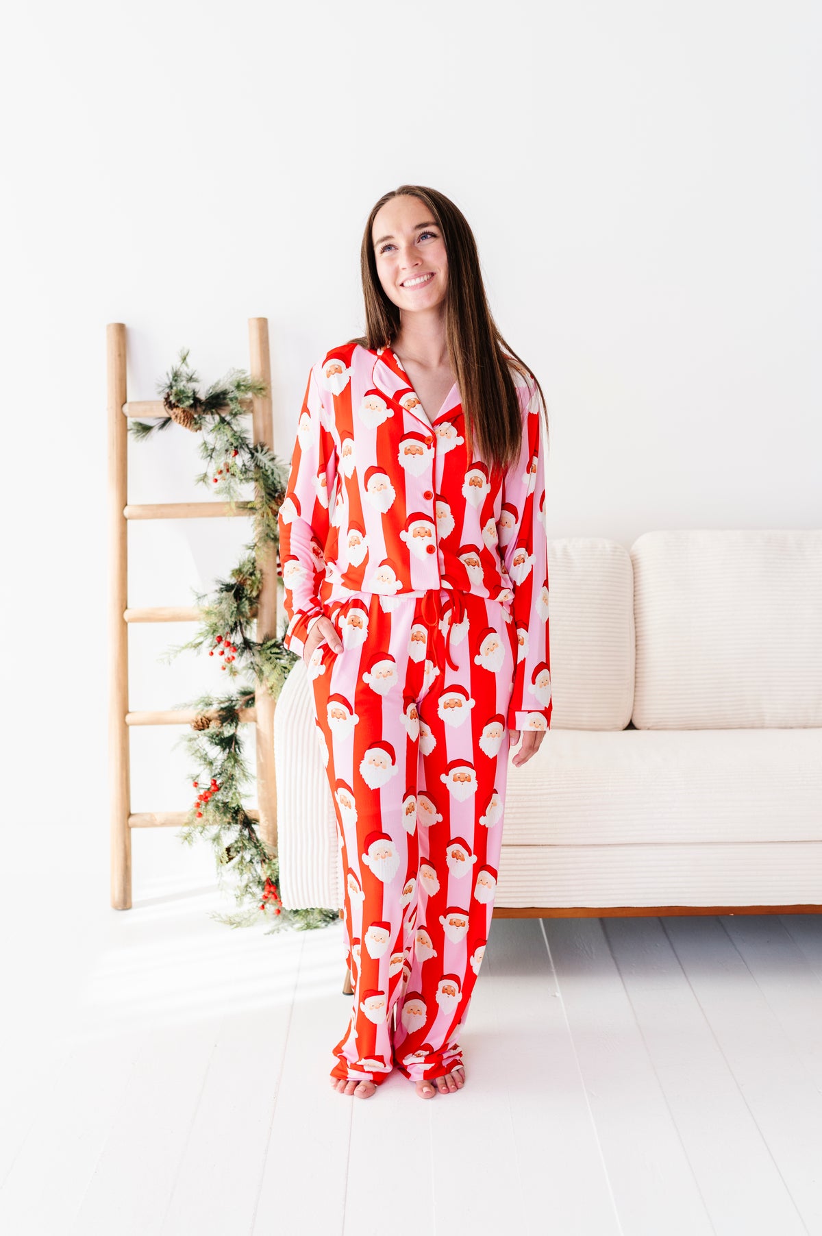 A woman stands in a bright white room wearing a red and white Santa Claus patterned bamboo pajama set. To her left is a wooden ladder decorated with green pine garlands, and behind her is a cream-colored sofa.