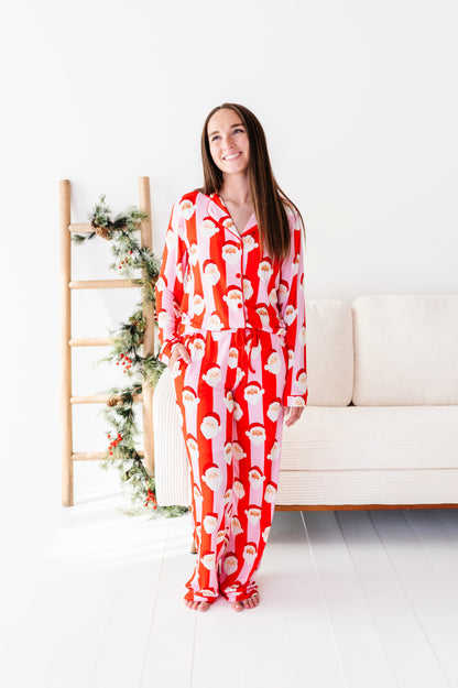 A woman stands in a bright white room wearing a red and white Santa Claus patterned bamboo pajama set. To her left is a wooden ladder decorated with green pine garlands, and behind her is a cream-colored sofa.
