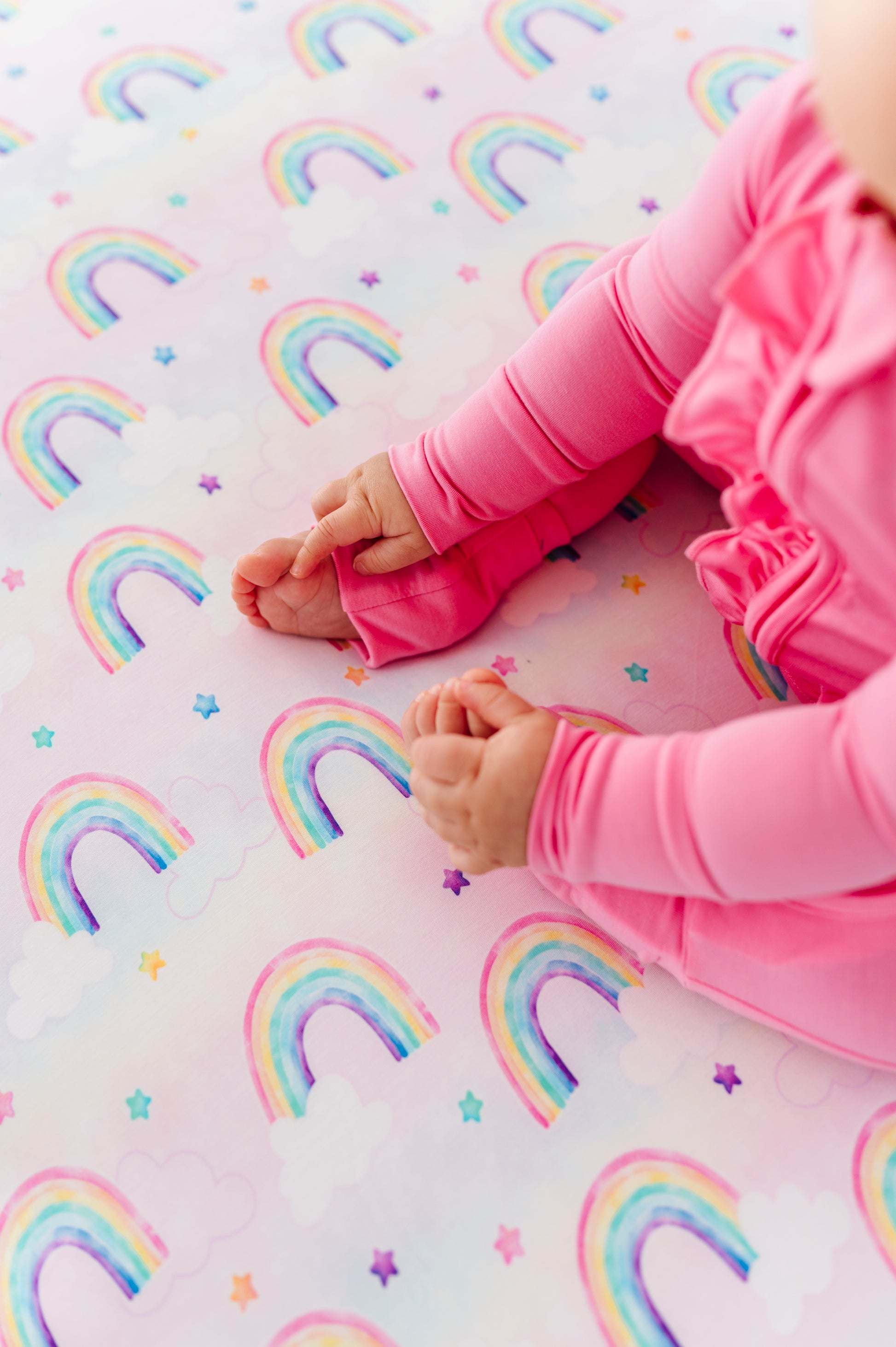Baby girl wearing a bamboo zip romper and sitting on a bamboo crib sheet featuring watercolor rainbow print 