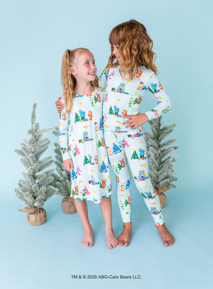 Two young girls wearing matching long-sleeve pajamas with a colorful Christmas tree and snowman print, standing barefoot against a light blue background with small potted trees.