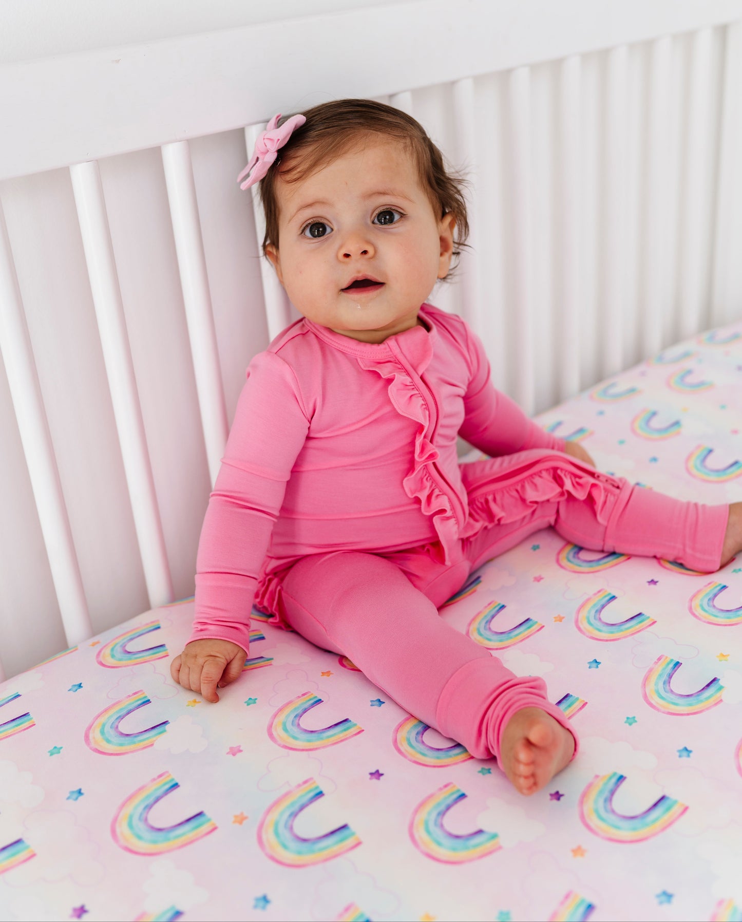 Baby in pink bamboo convertible romper sitting on a crib with rainbow-patterned bedding