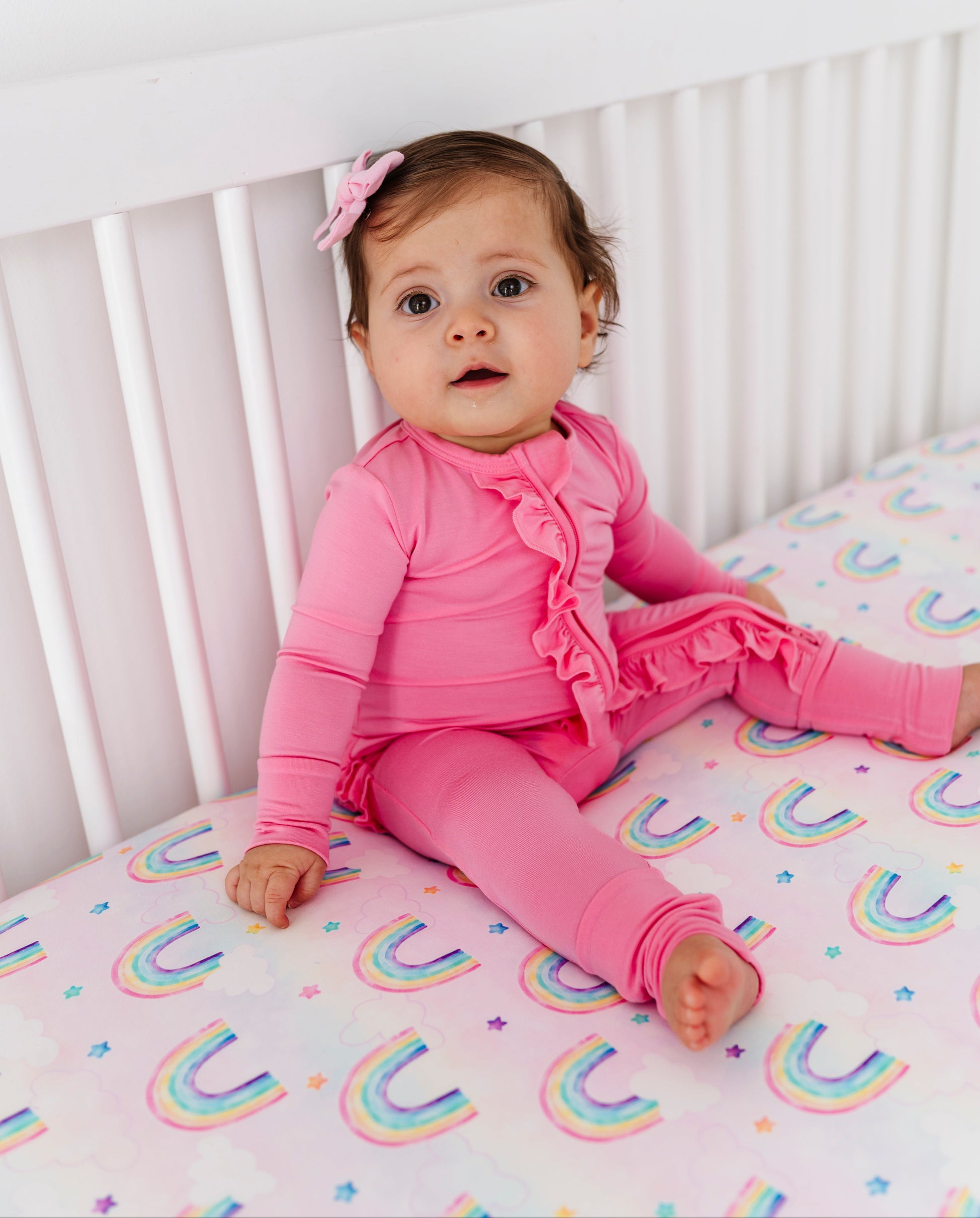 Baby in pink bamboo convertible romper sitting on a crib with rainbow-patterned bedding