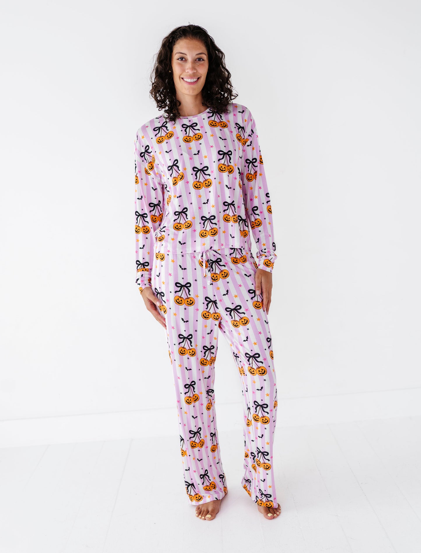 A woman modeling a light pink long-sleeve pajama set with a black cat and orange pumpkin pattern, standing against a plain white studio background.