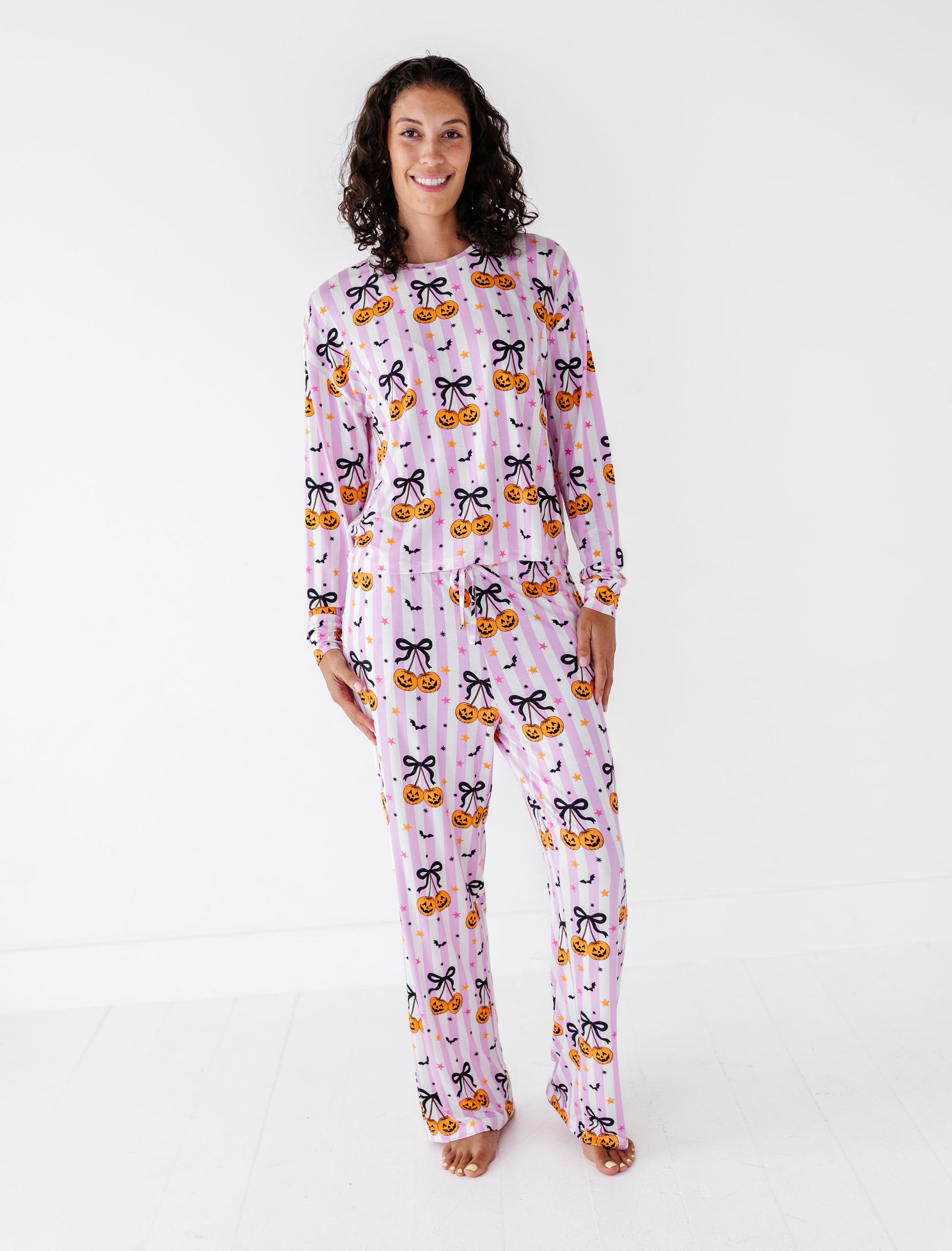 A woman modeling a light pink long-sleeve pajama set with a black cat and orange pumpkin pattern, standing against a plain white studio background.