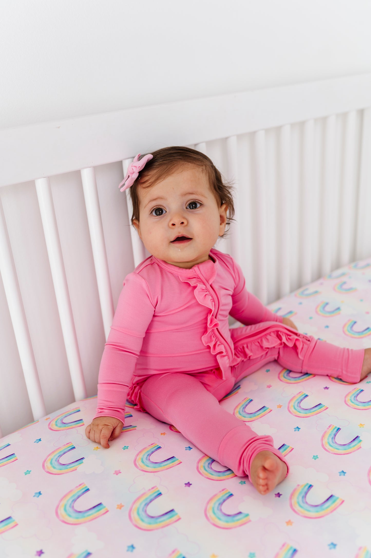 Baby in pink bamboo zip romper sitting on a crib with rainbow-patterned bamboo crib sheet