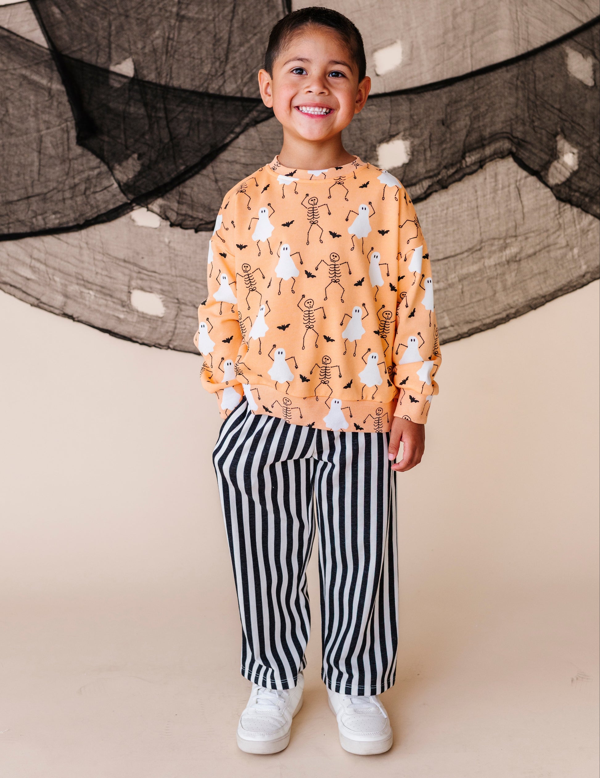 Child wearing a patterned sweatshirt and striped pants standing against a neutral background