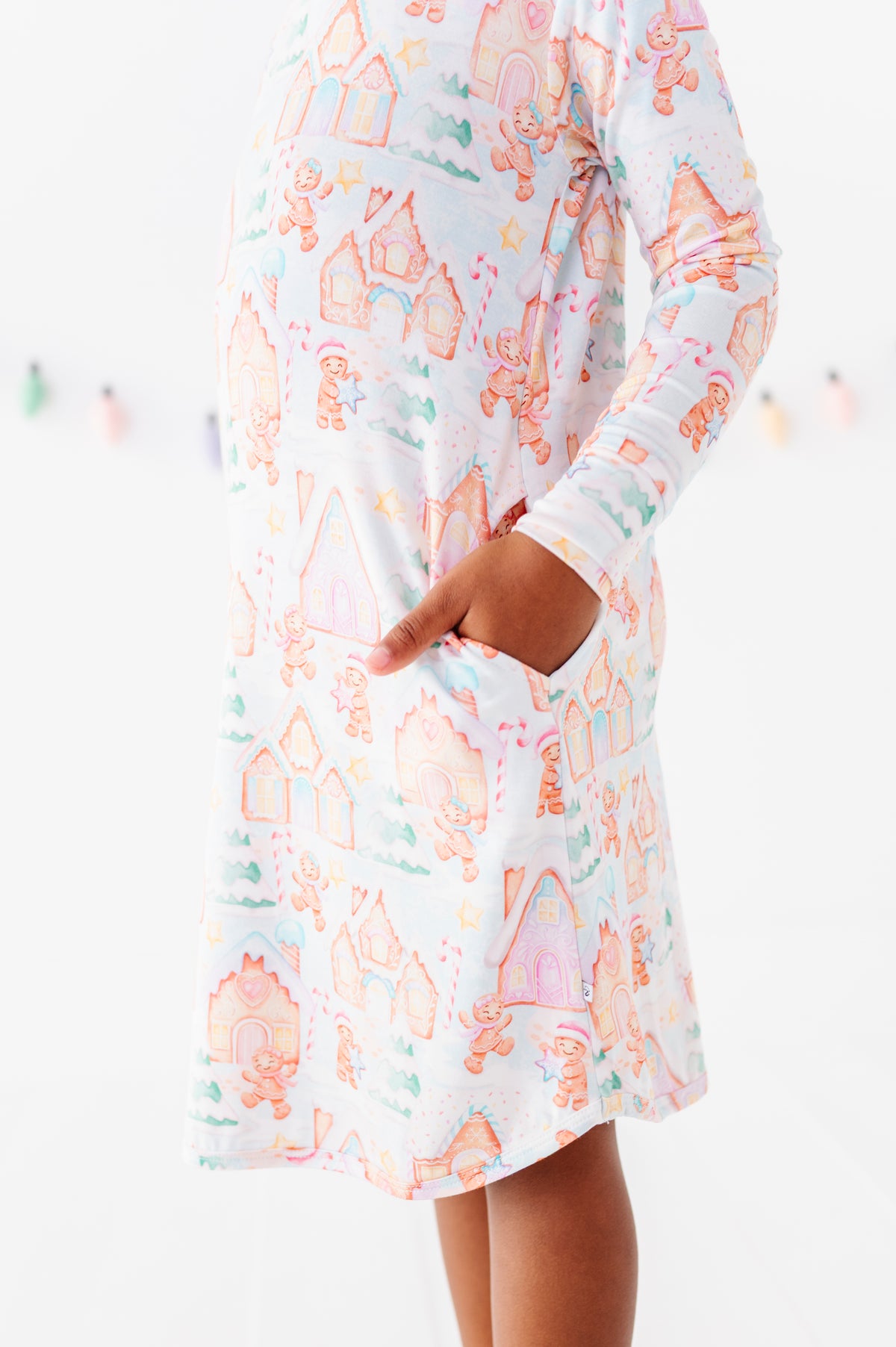 A close-up detail shot of a little girl wearing a Christmas gingerbread-themed bamboo nightgown, standing in a bright white room 