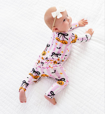 A baby lying on a white quilted blanket, wearing a pink long-sleeve footed pajama with a pumpkin and bow pattern, and a white bow headband.