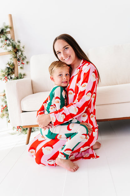 A woman and a young boy sitting on a white floor in front of a cream sofa, wearing matching Christmas-themed bamboo pajamas with Santa Claus patterns in red, green, and white.