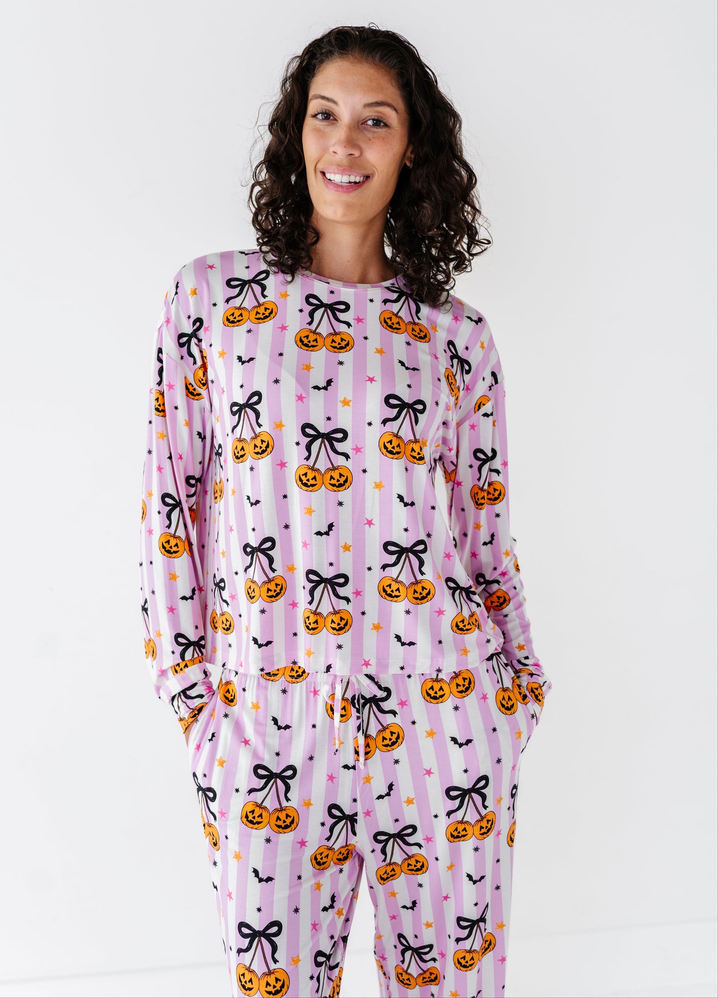 A woman modeling a long-sleeve pajama set with a pink and white striped Halloween pattern featuring pumpkins, black bows, and bats against a plain white background.