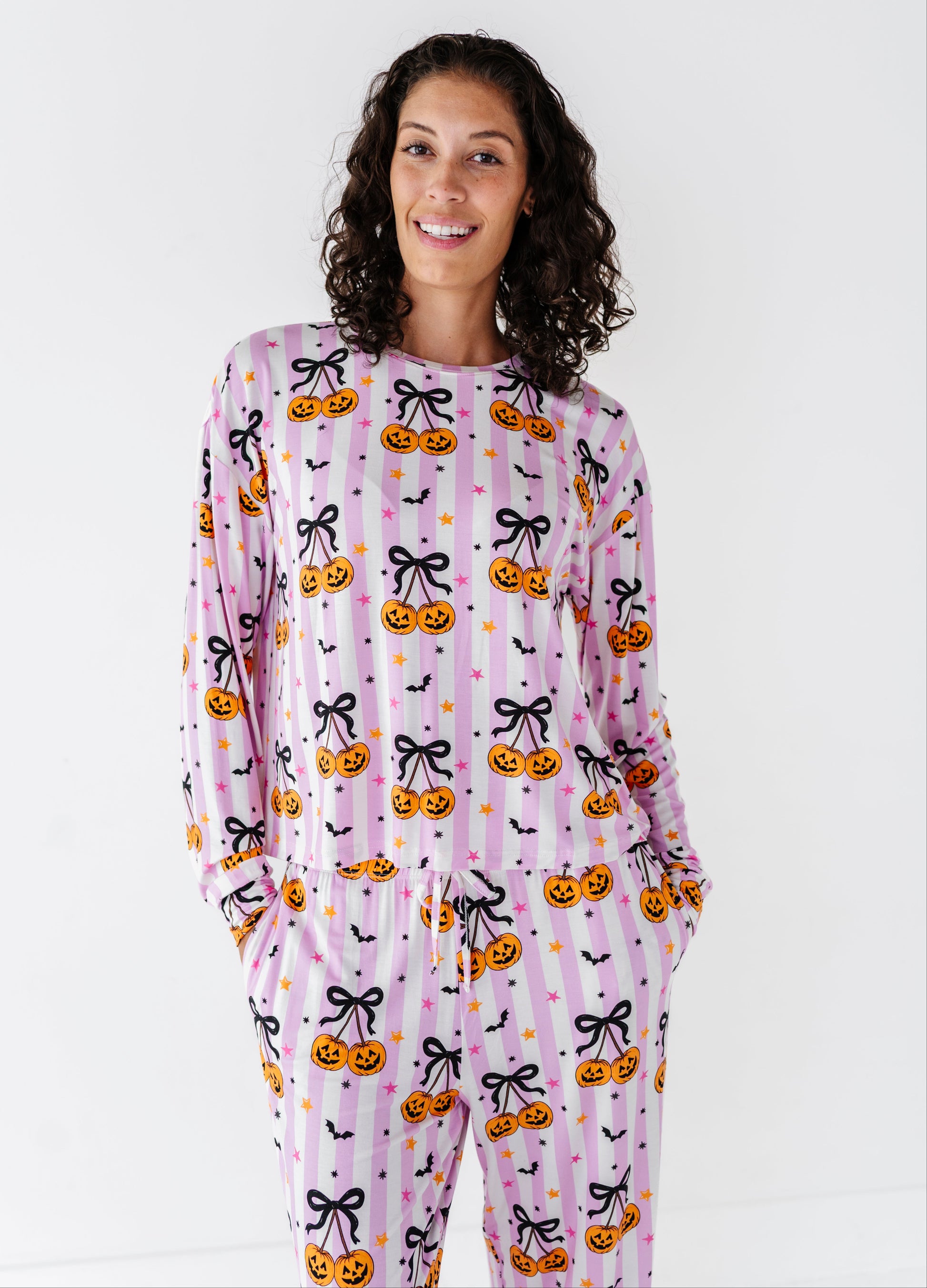 A woman modeling a long-sleeve pajama set with a pink and white striped Halloween pattern featuring pumpkins, black bows, and bats against a plain white background.