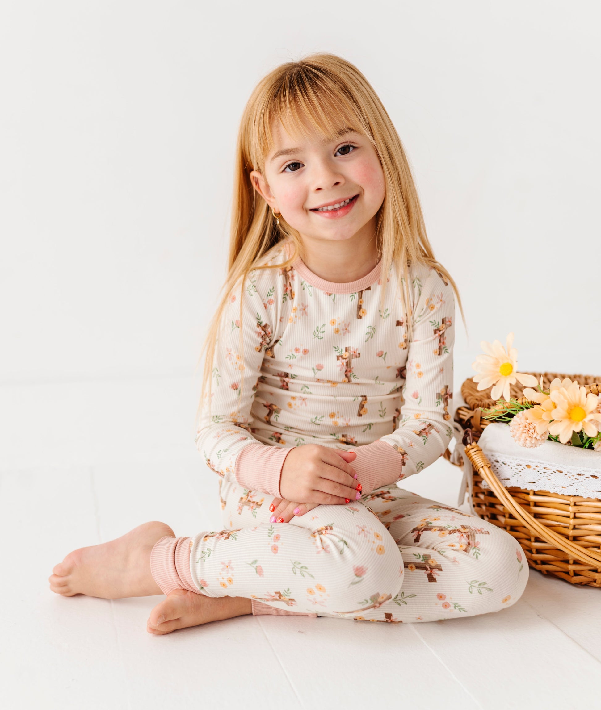 Young girl in a floral bamboo pajamas sitting on a white floor with a basket and flowers in the background.