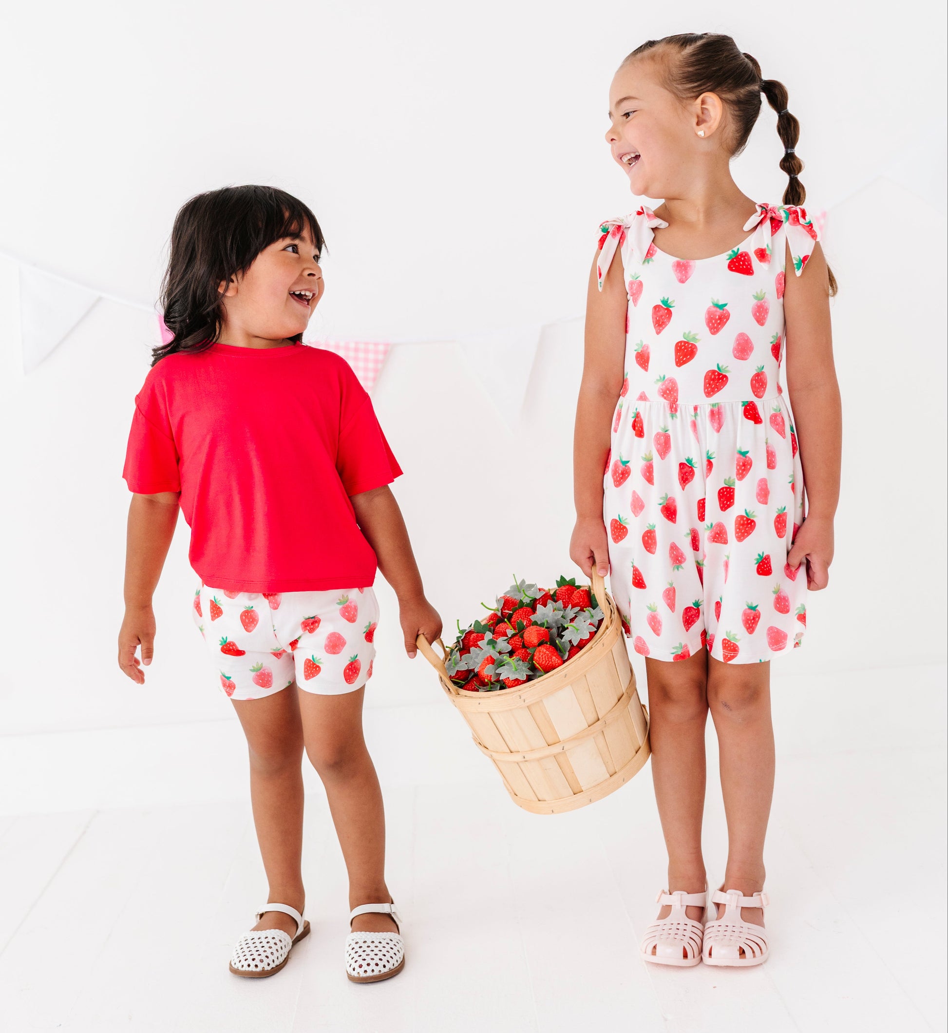 Two children in matching strawberry-themed outfits holding a basket of strawberries on a white background