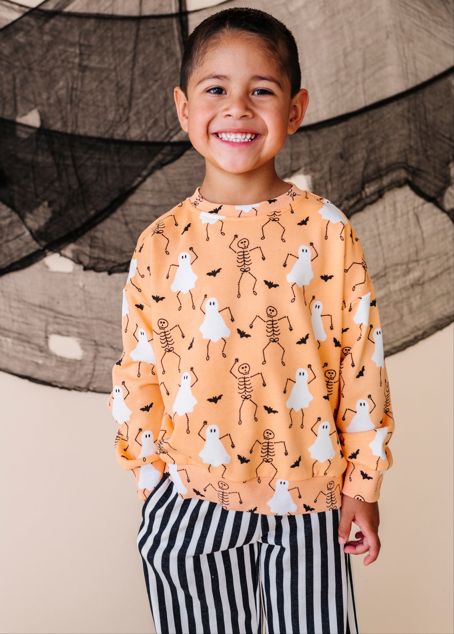 Child wearing a Halloween-themed sweatshirt with ghosts and skeletons in front of a black spider web.