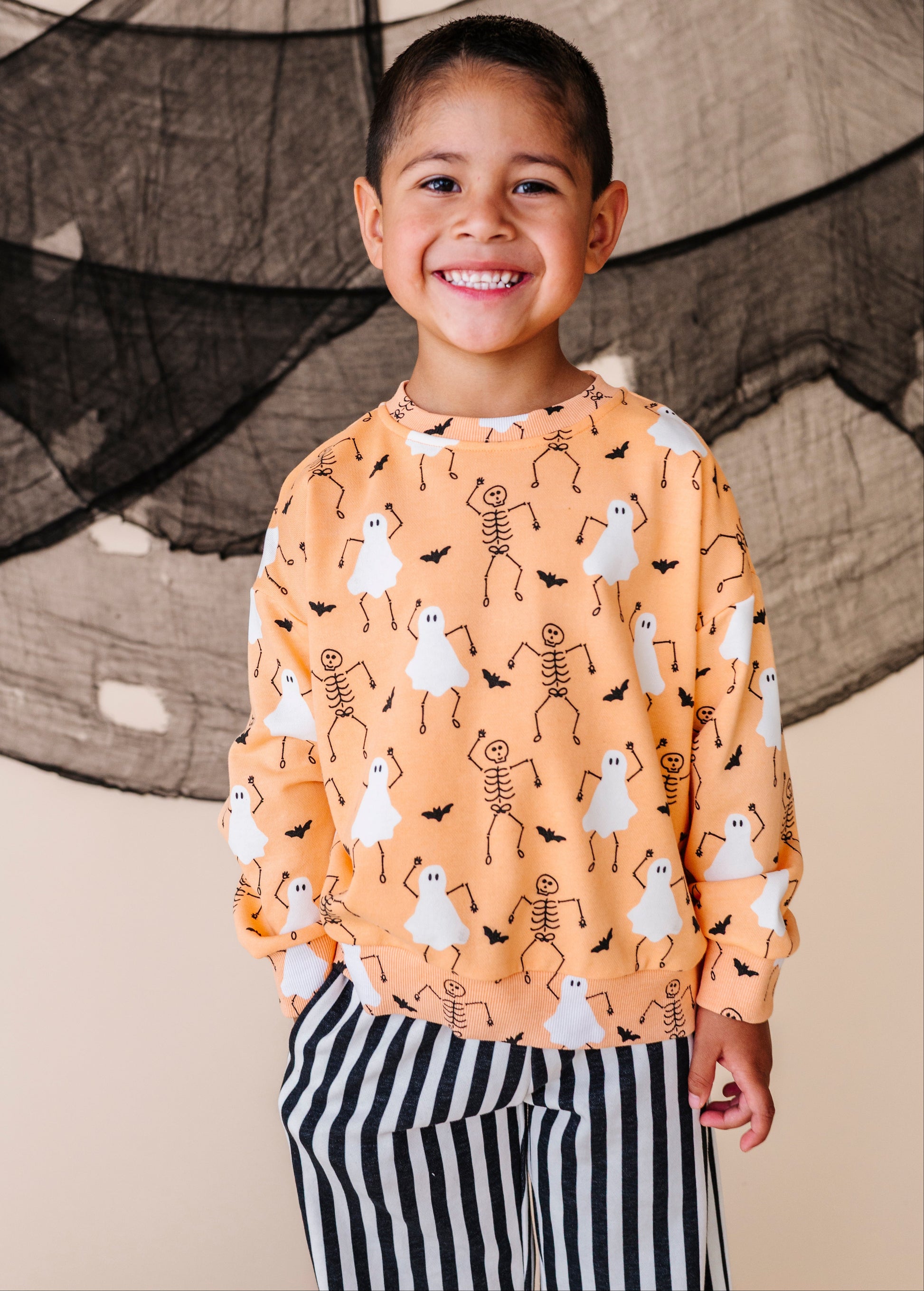 Child wearing a Halloween-themed sweatshirt with ghosts and skeletons in front of a black spider web.