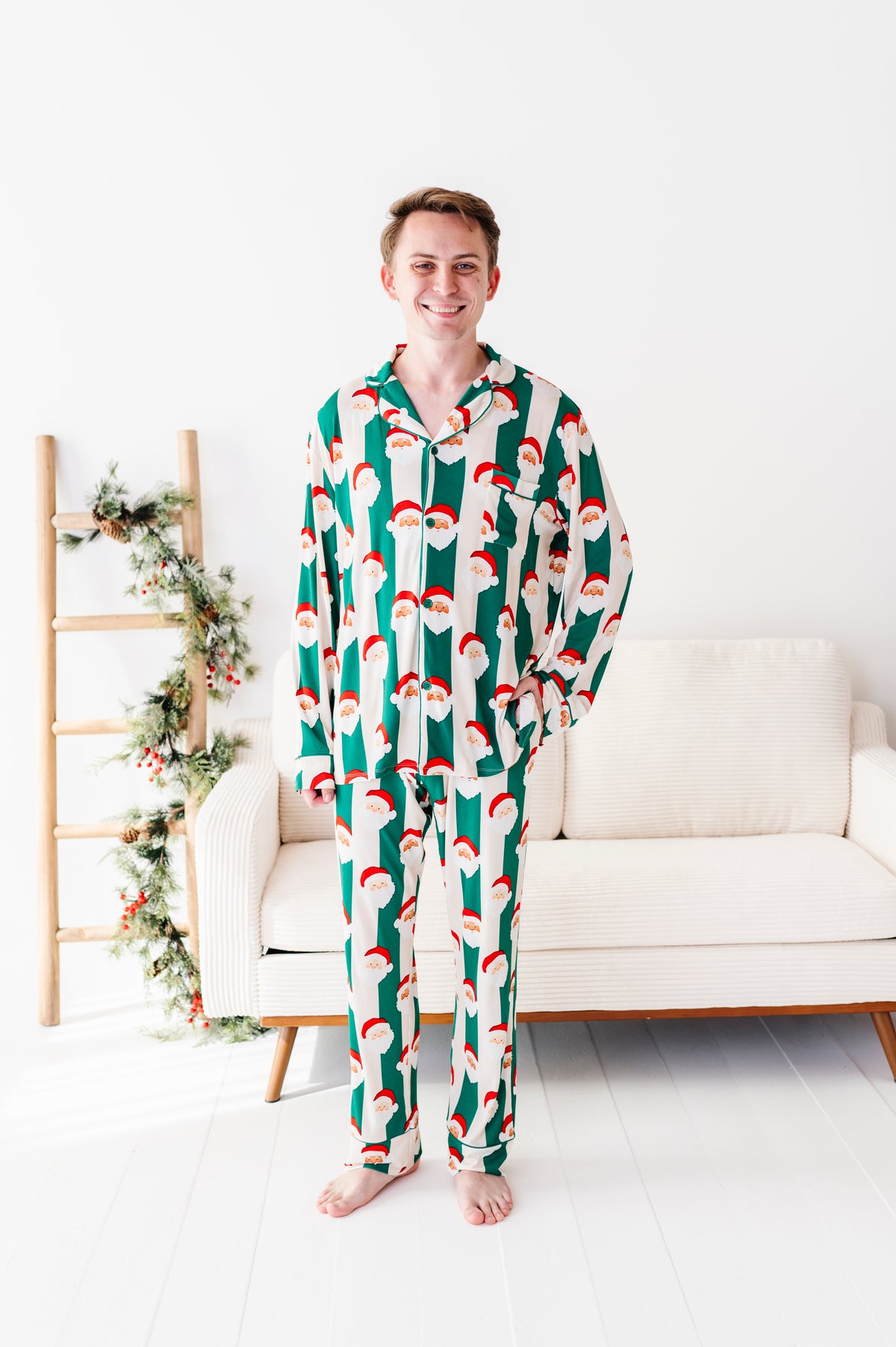 A man wearing a green and white Santa Claus patterned bamboo pajama set stands barefoot in a bright, modern room with a white sofa and a wooden ladder decorated with greenery.