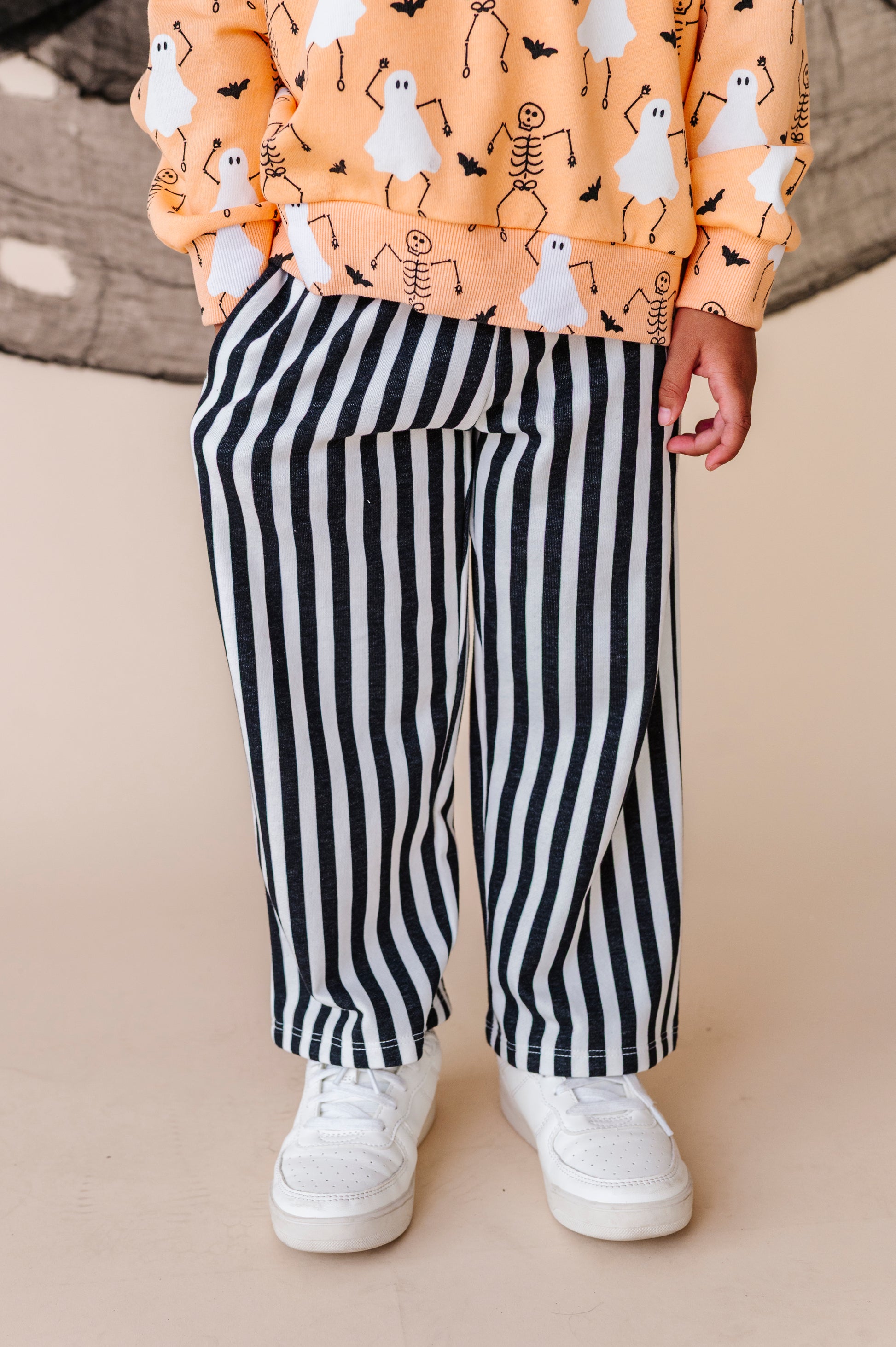 Child wearing a Halloween-themed shirt with ghosts and skeletons, black and white striped pants, and white sneakers on a beige background.