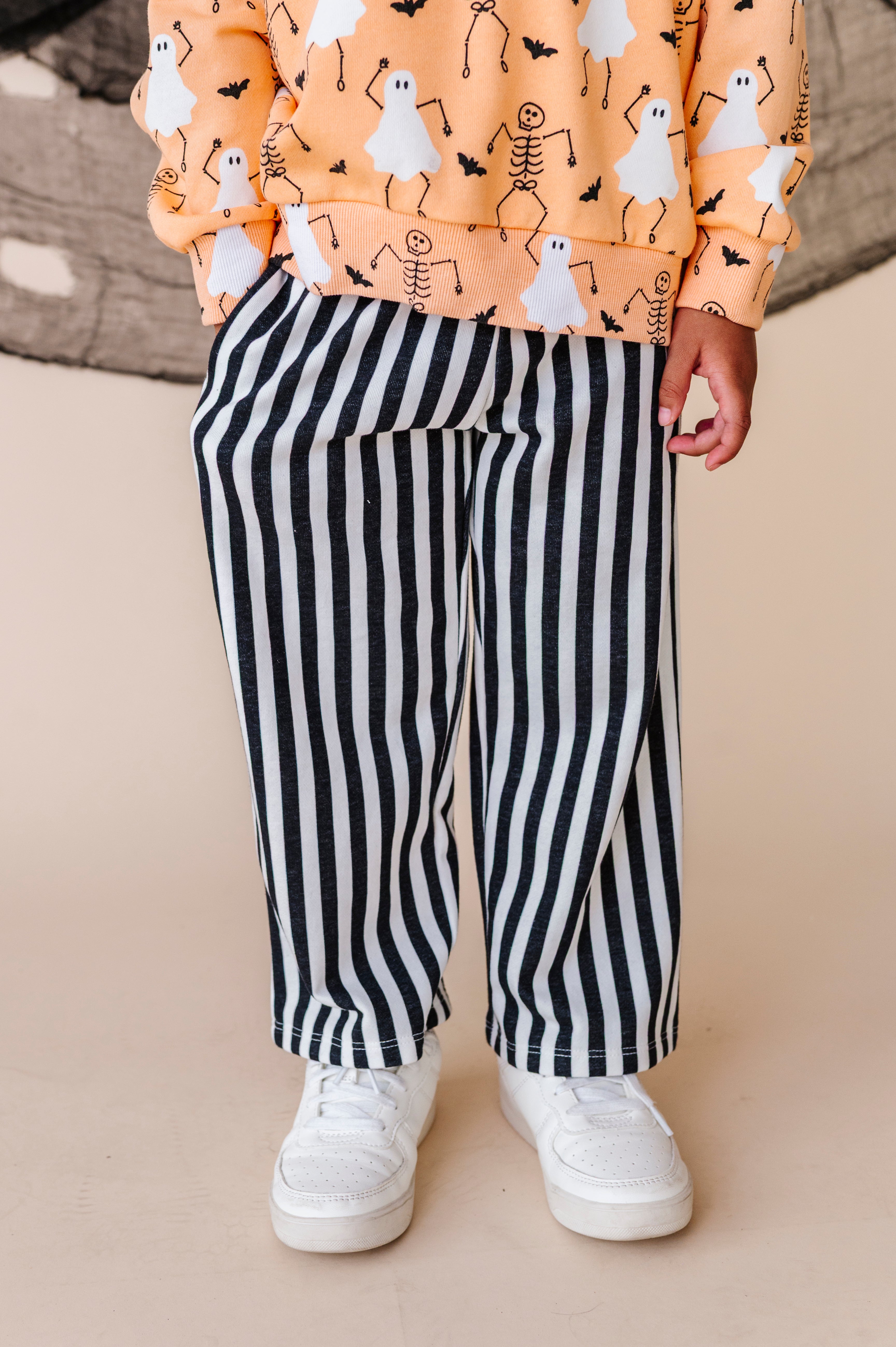 Child wearing a Halloween-themed shirt with ghosts and skeletons, black and white striped pants, and white sneakers on a beige background.