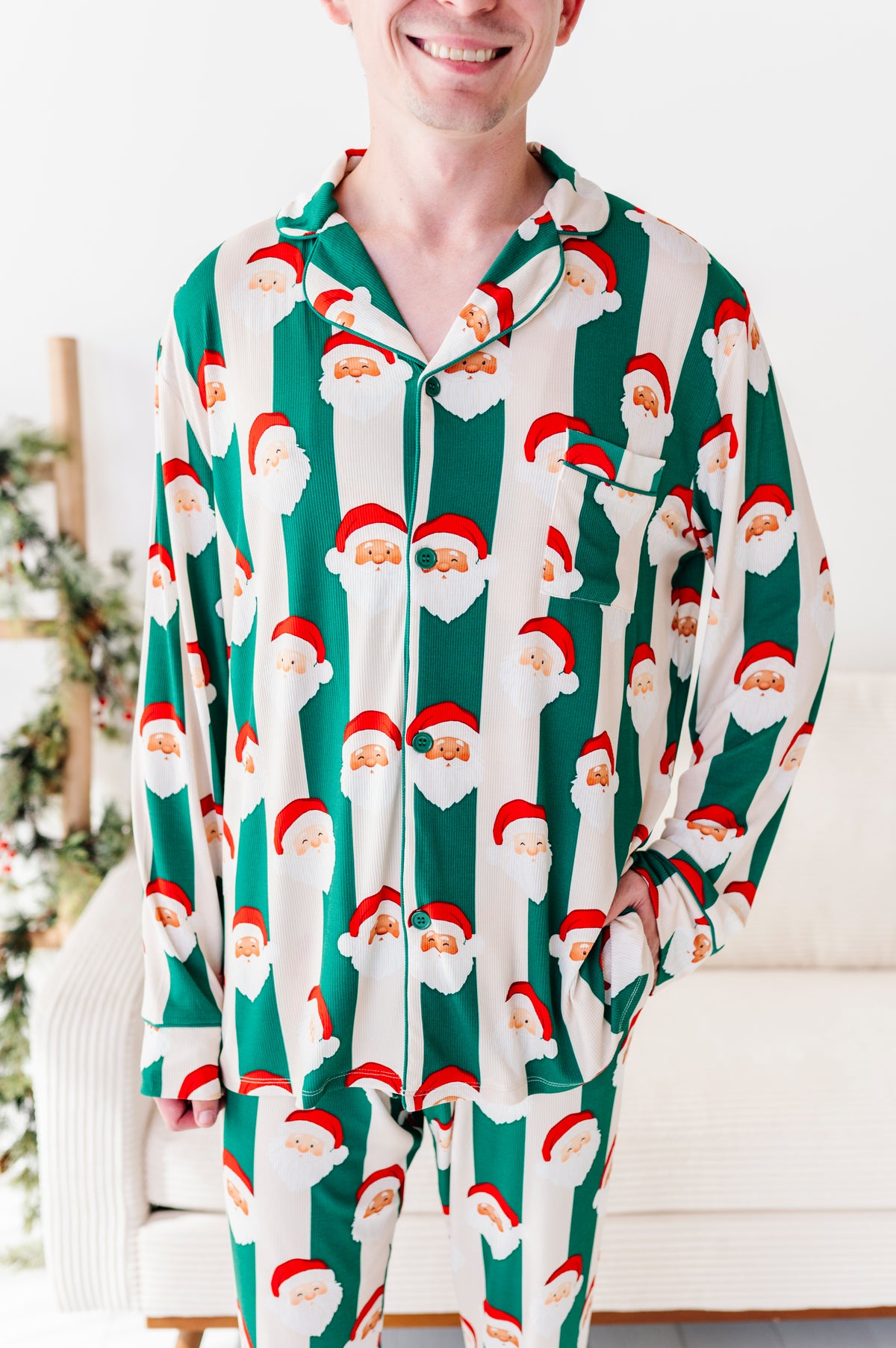 A man wearing a long-sleeve bamboo pajama set with a green and white striped pattern featuring Santa Claus faces, standing in a brightly lit indoor setting.