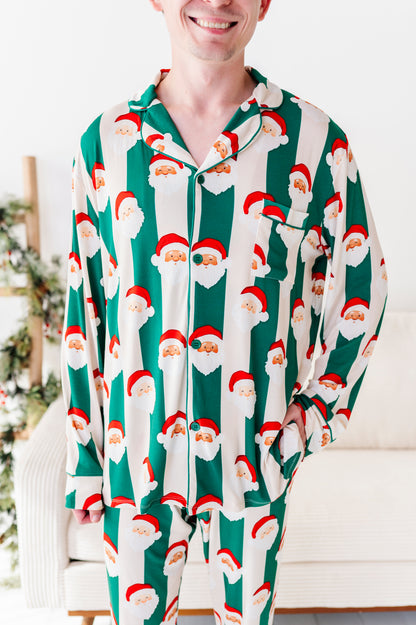 A man wearing a long-sleeve bamboo pajama set with a green and white striped pattern featuring Santa Claus faces, standing in a brightly lit indoor setting.