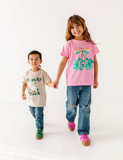 Two children holding hands, one wearing a pink shirt with a palm tree design and the other wearing a beige shirt with text.