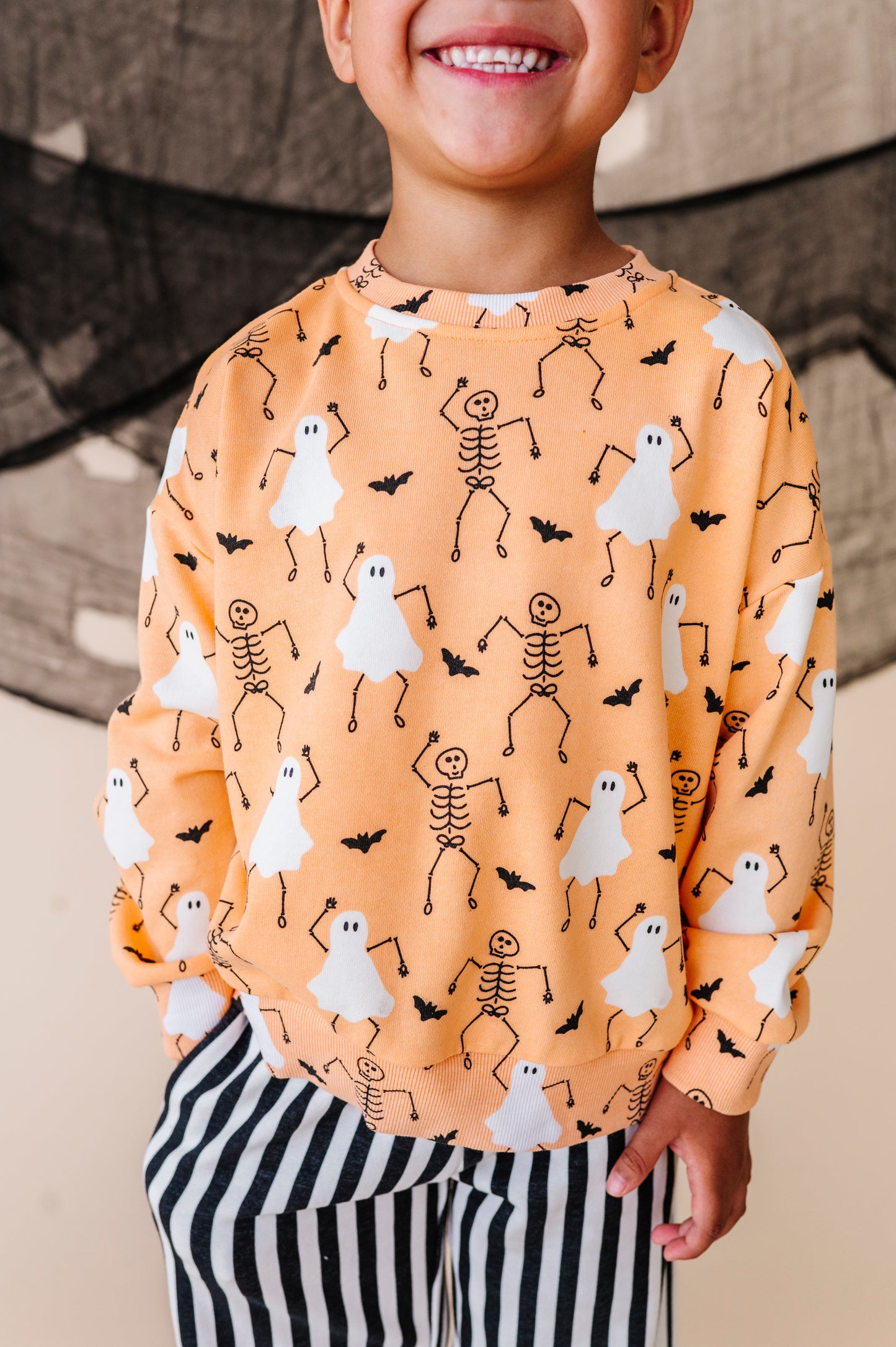 Child wearing a Halloween-themed sweatshirt with ghosts, skeletons, and bats on an orange background.