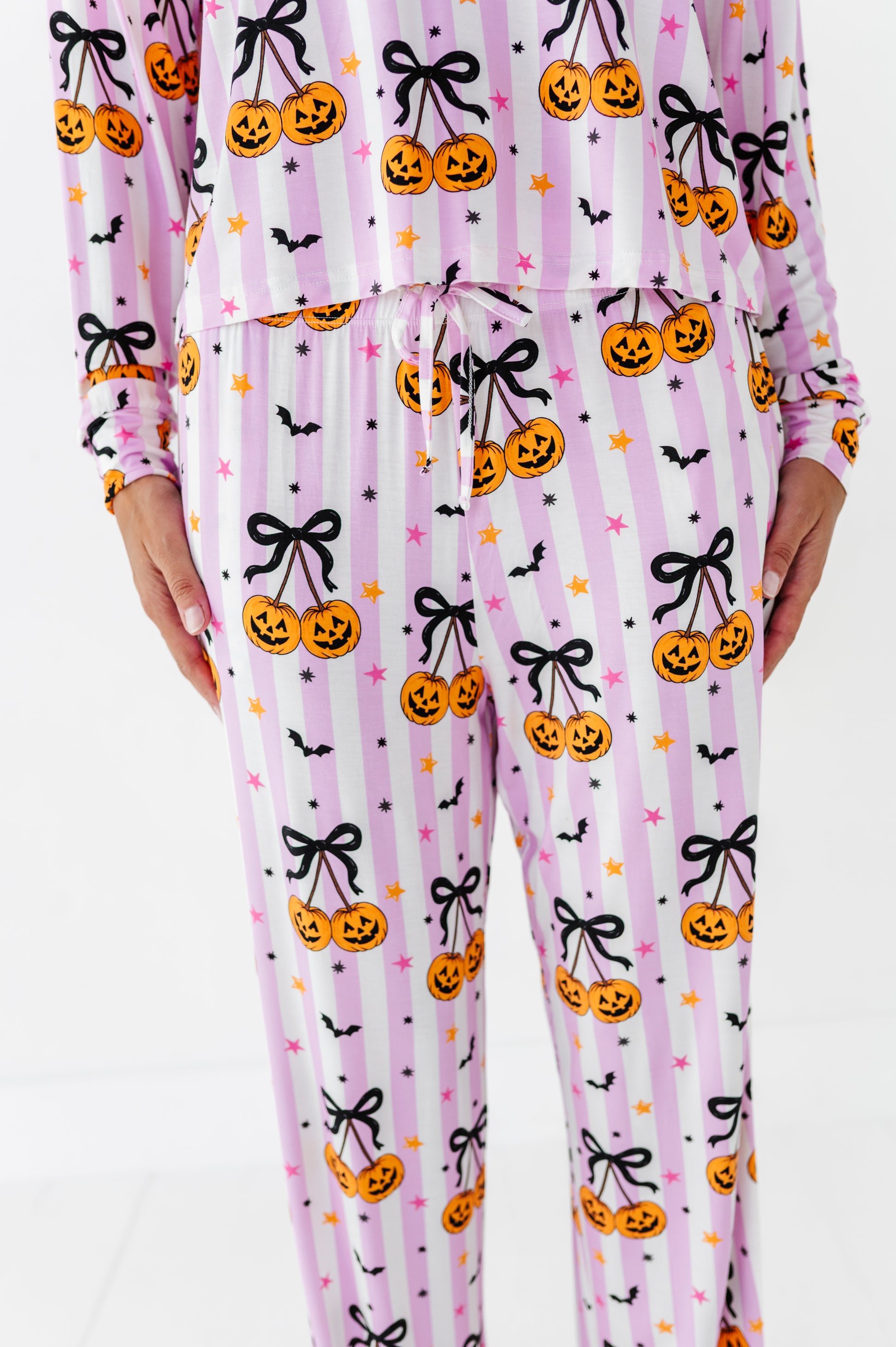 A person wearing a matching long-sleeve pajama top and pants set with a pink and white vertical stripe pattern, decorated with orange jack-o'-lanterns, black bows, black bats, and small pink and orange stars.